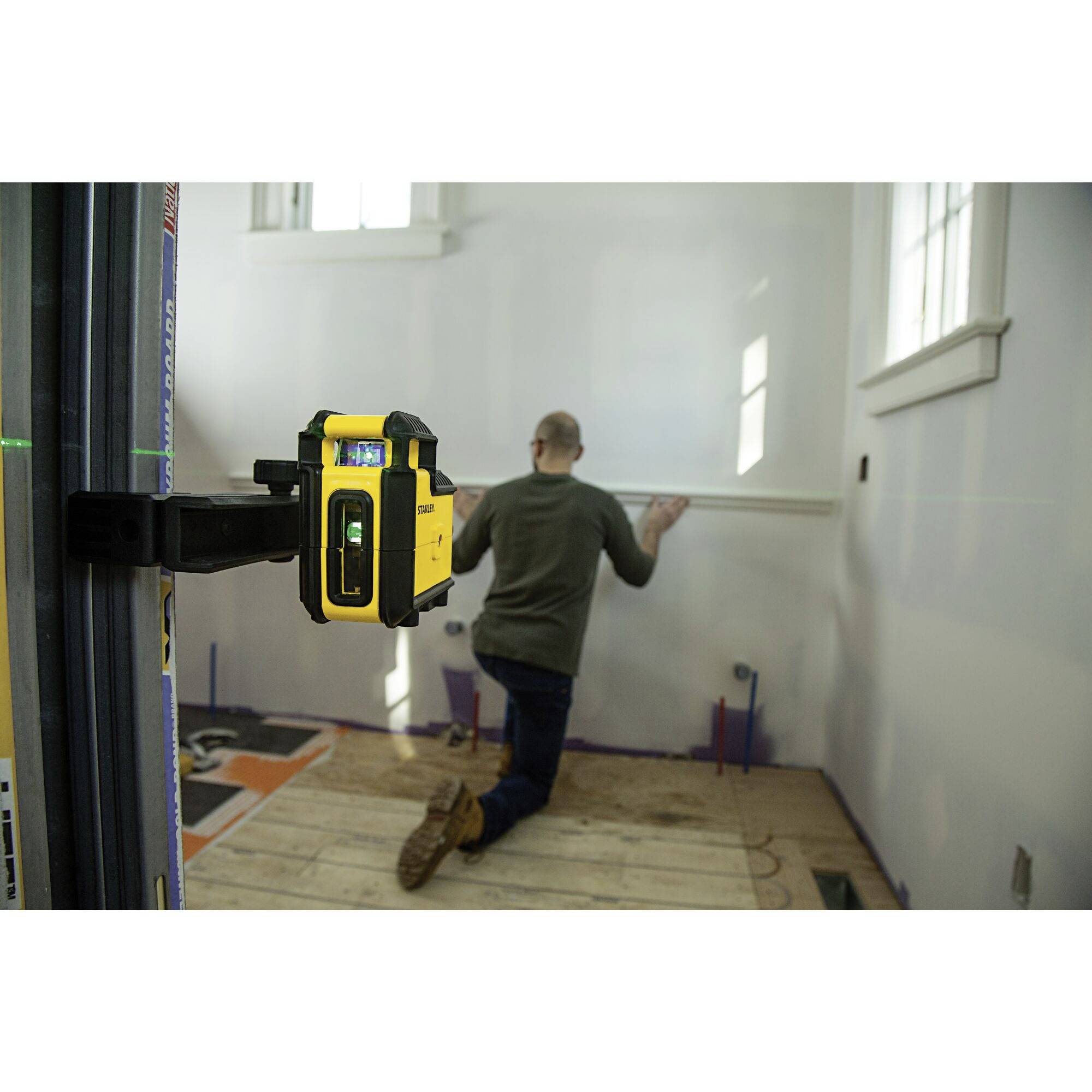 A man is installing a strip on the wall using a yellow laser tool in a white room.