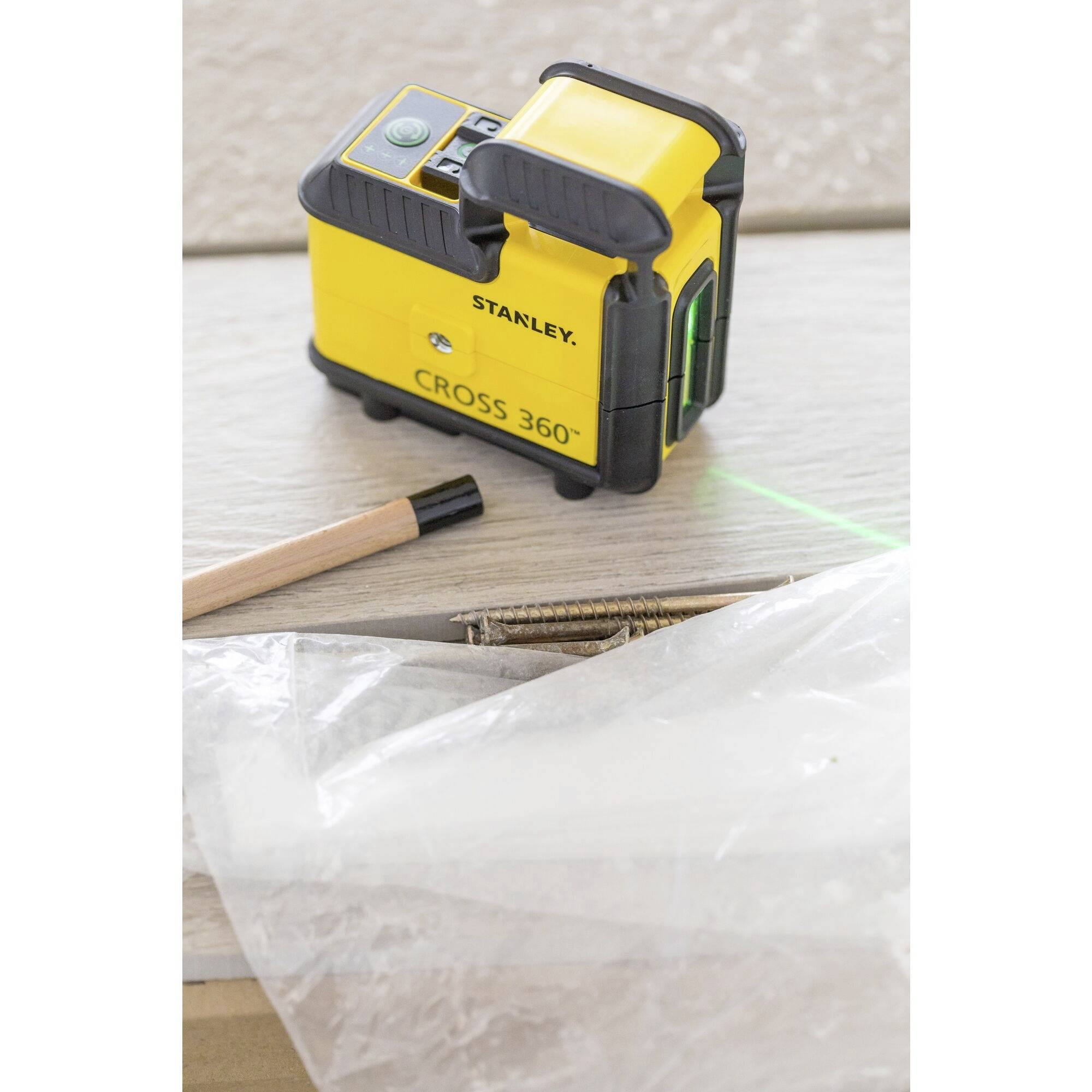 A yellow Stanley Cross 360° laser level tool on a wooden table, surrounded by plastic sheeting and a pencil.