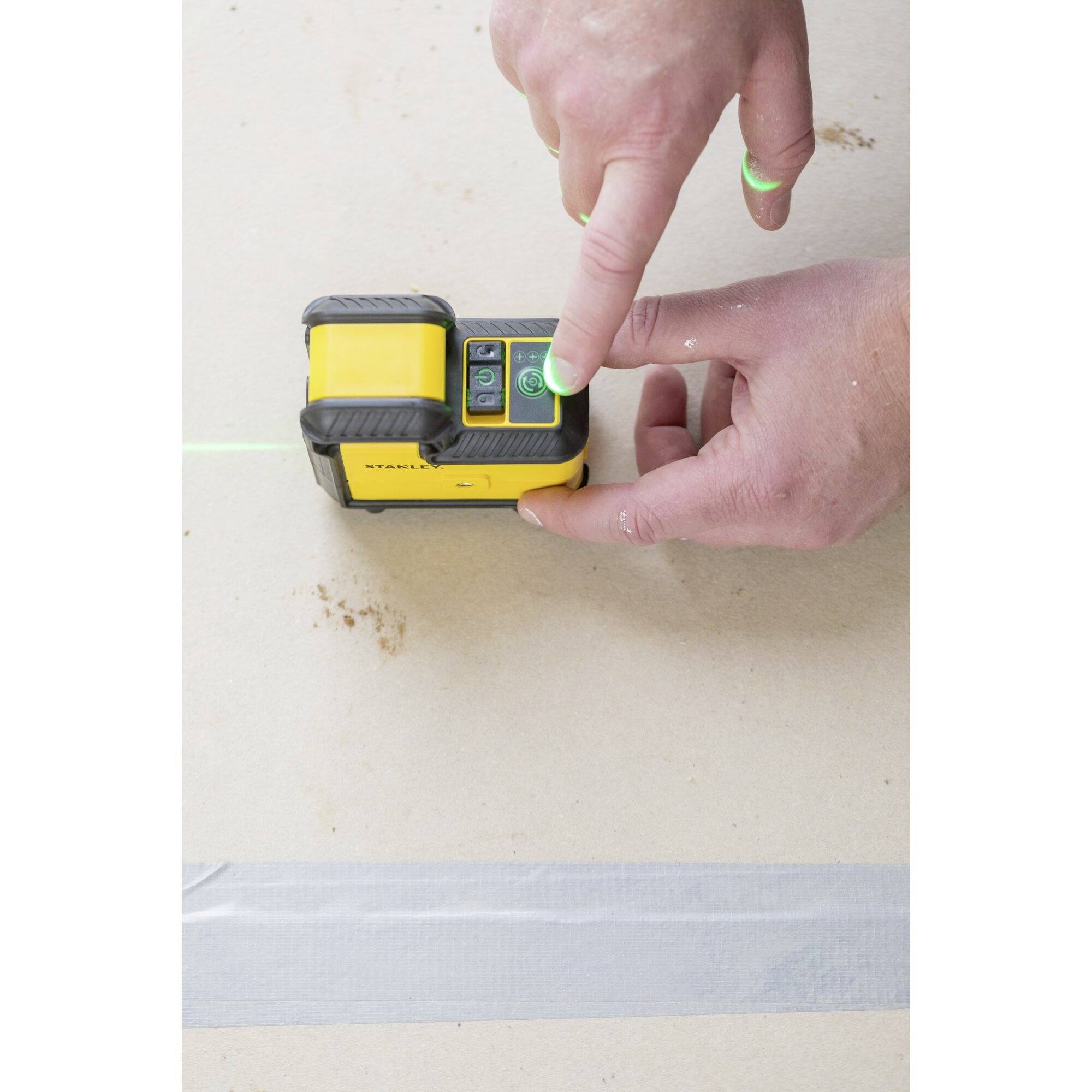 A person is operating a yellow laser levelling device on a table. A green laser beam is active, shining onto the surface.