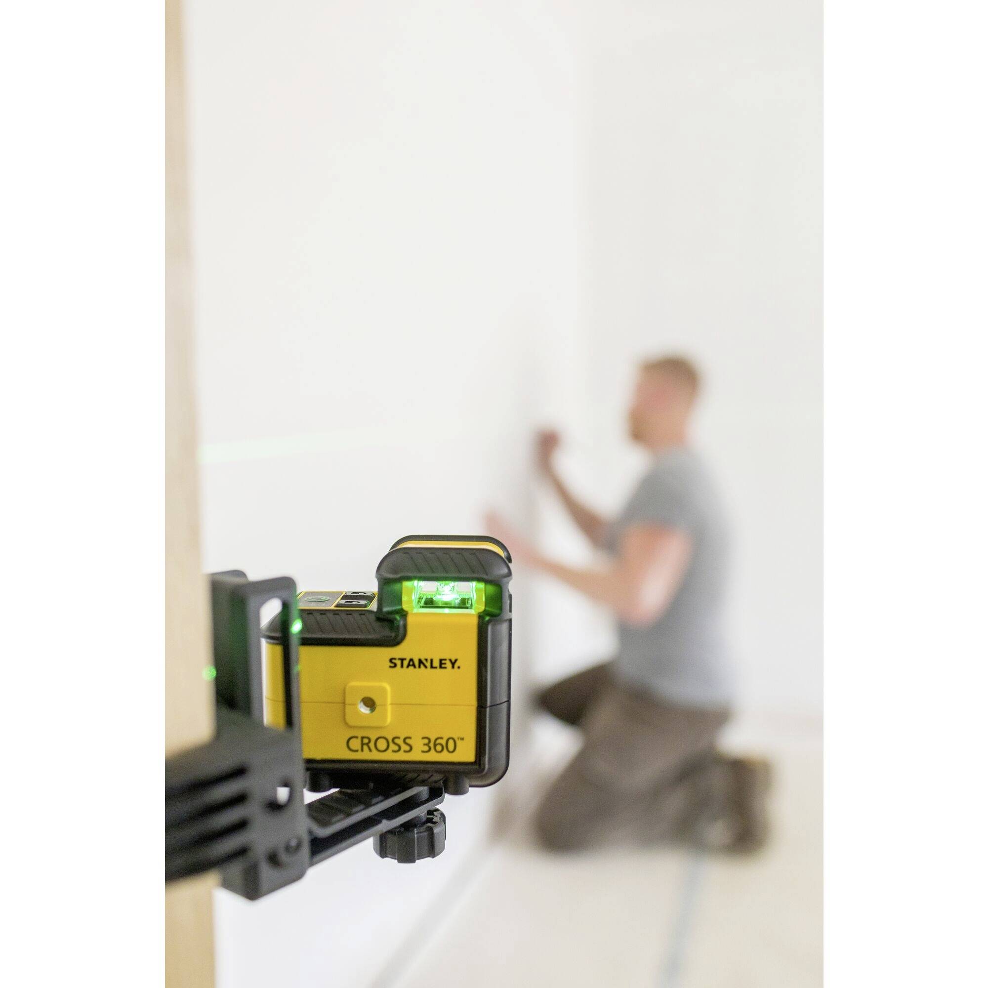 A man is kneeling in front of a wall and installing a laser levelling device marked 'Cross 360°' to set precise markings.