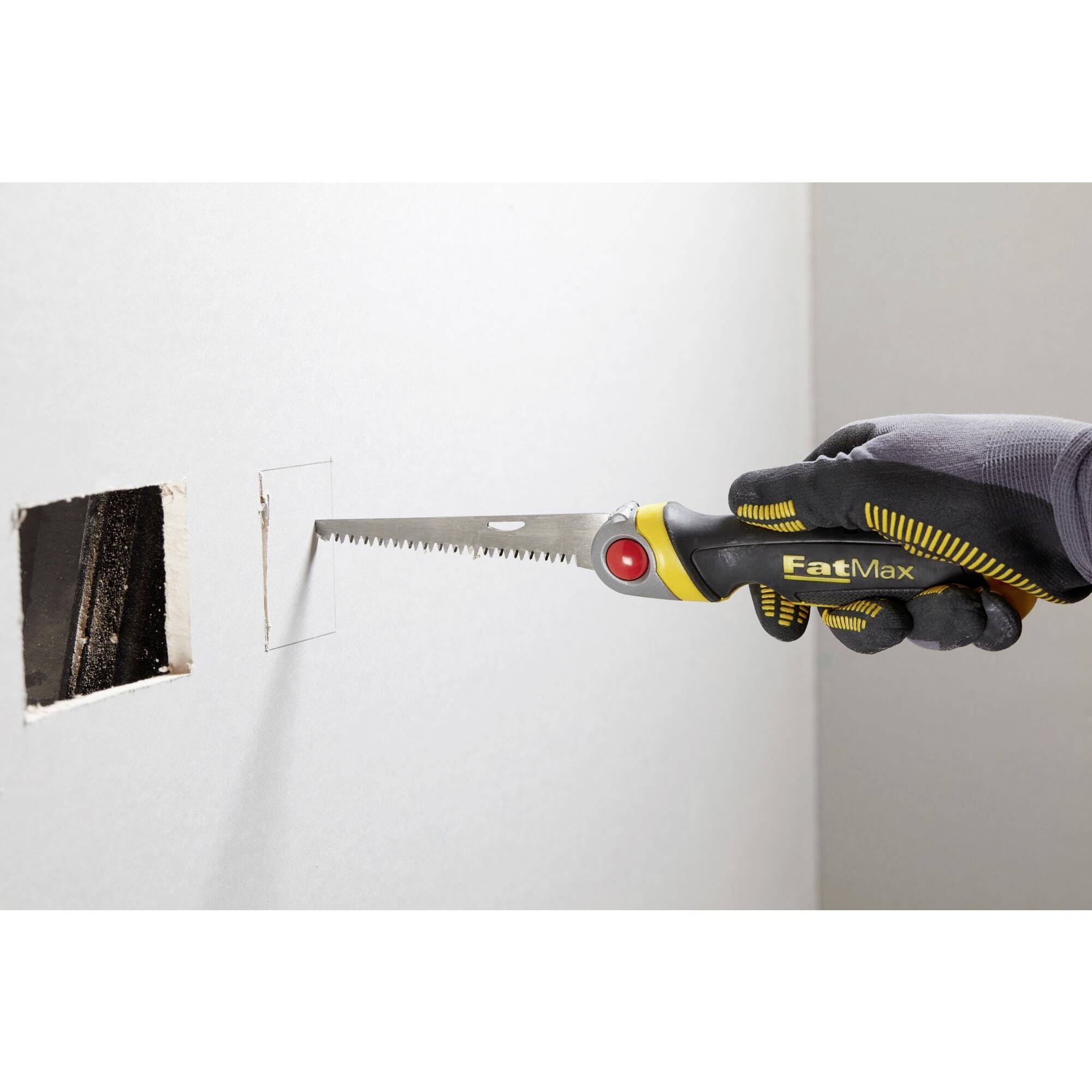 A hand is cutting a square hole in a wall using a handsaw. The saw has a yellow handle with a red insert.