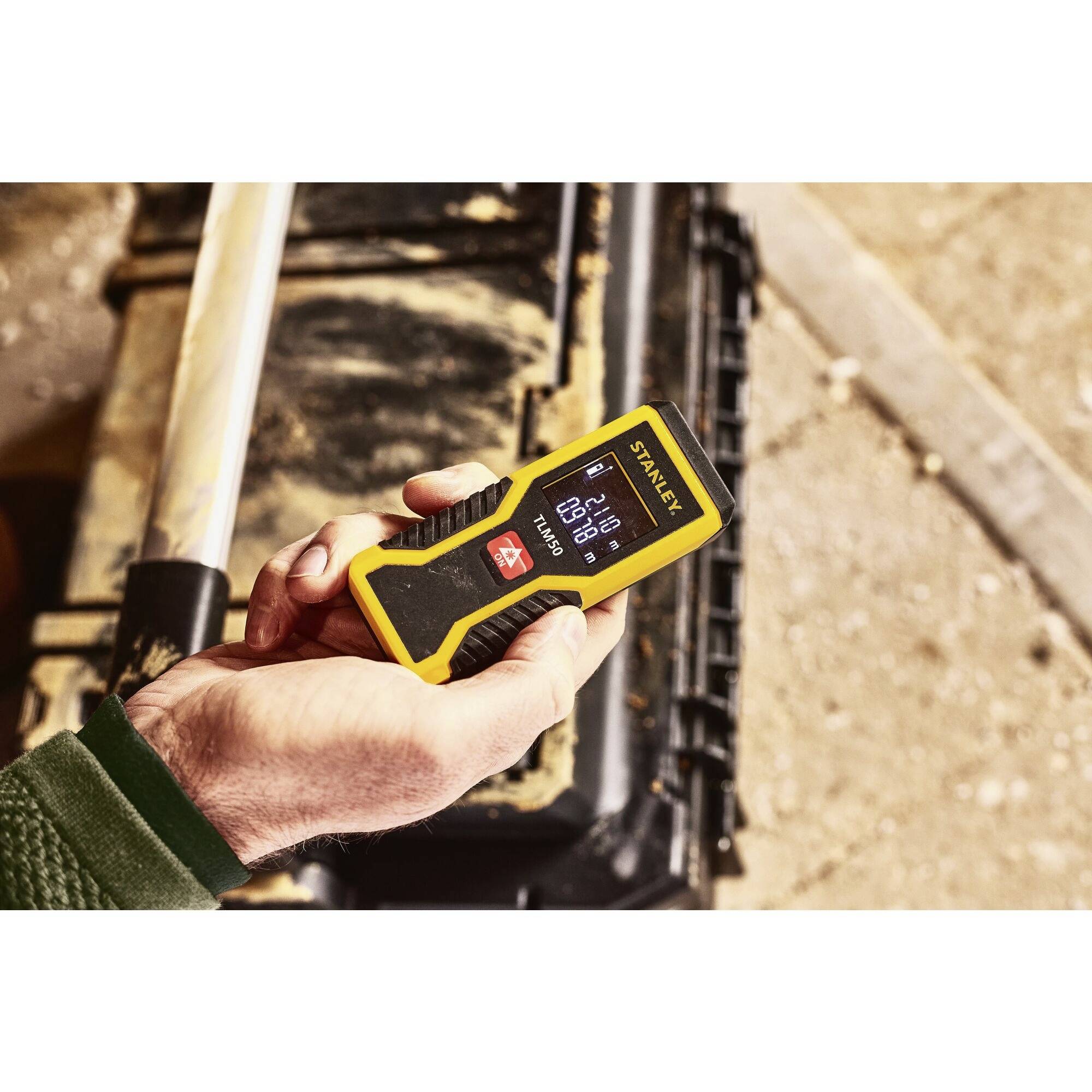 A hand is holding a digital yellow measuring device displaying 0.12 metres. A toolbox can be seen in the background.
