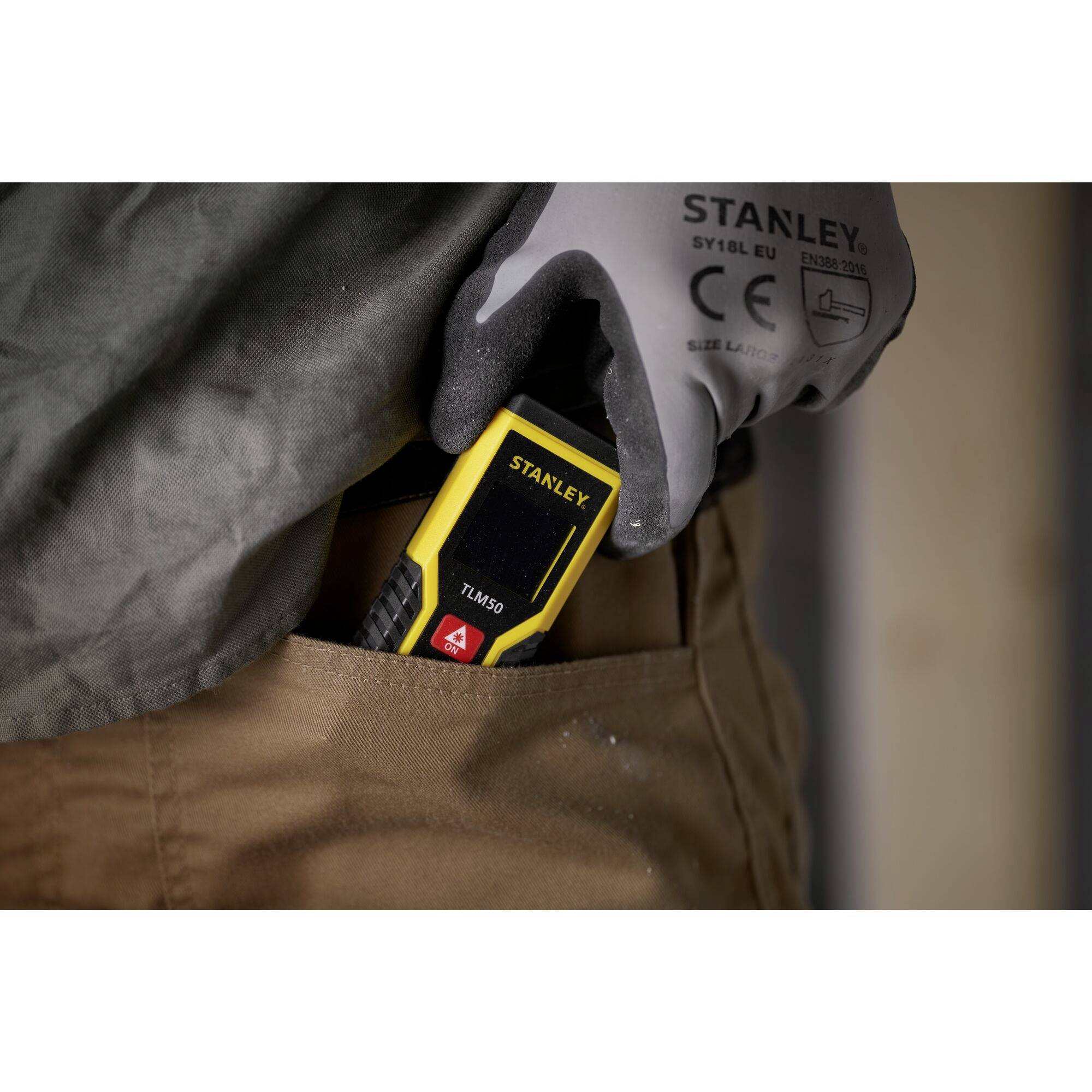 A person pulls out a yellow Stanley measuring device from their trouser or jacket pocket. They are wearing a grey glove.