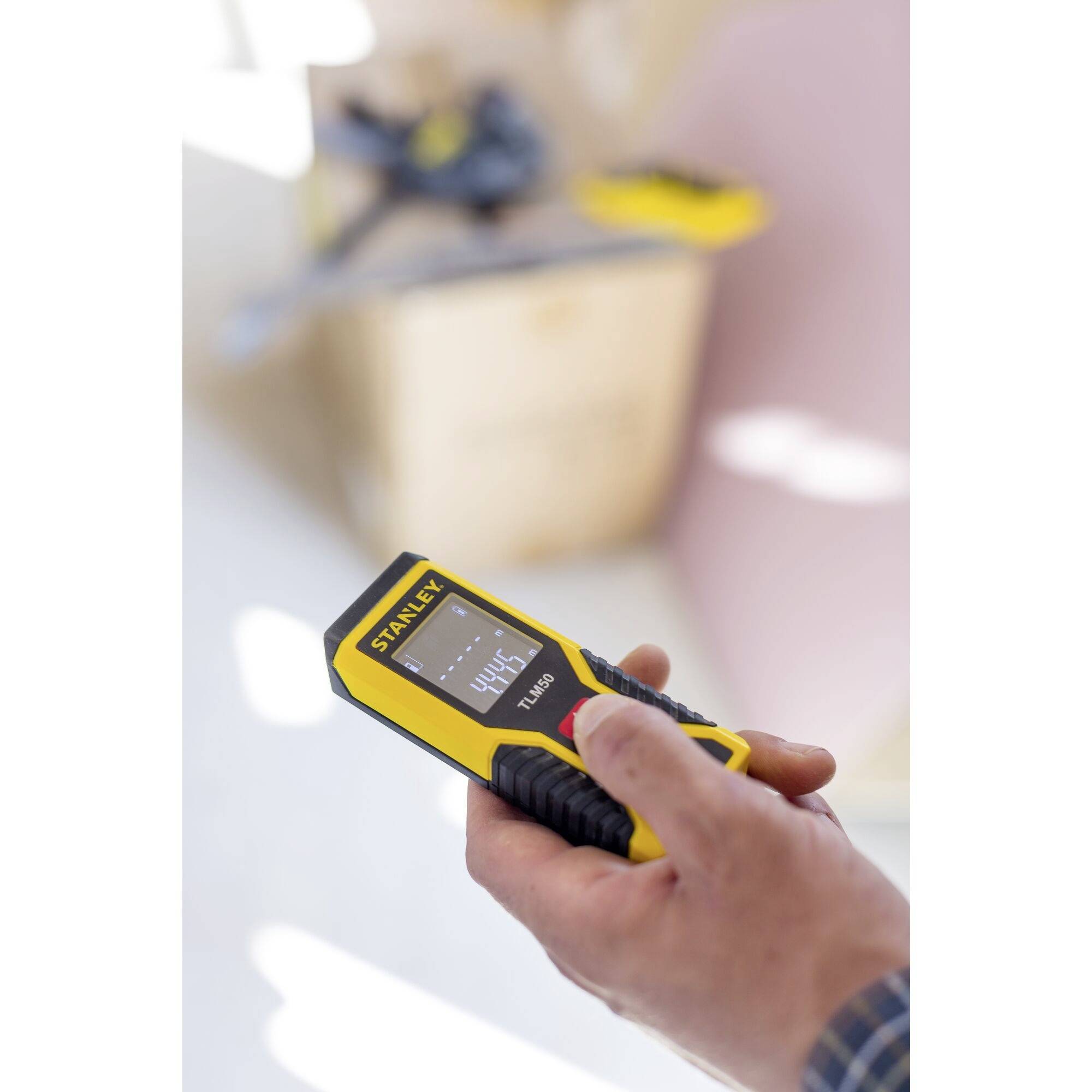 A hand is holding a yellow digital distance measurer by Staley, displaying a measurement distance of 444 mm. Blurred tools are visible in the background.