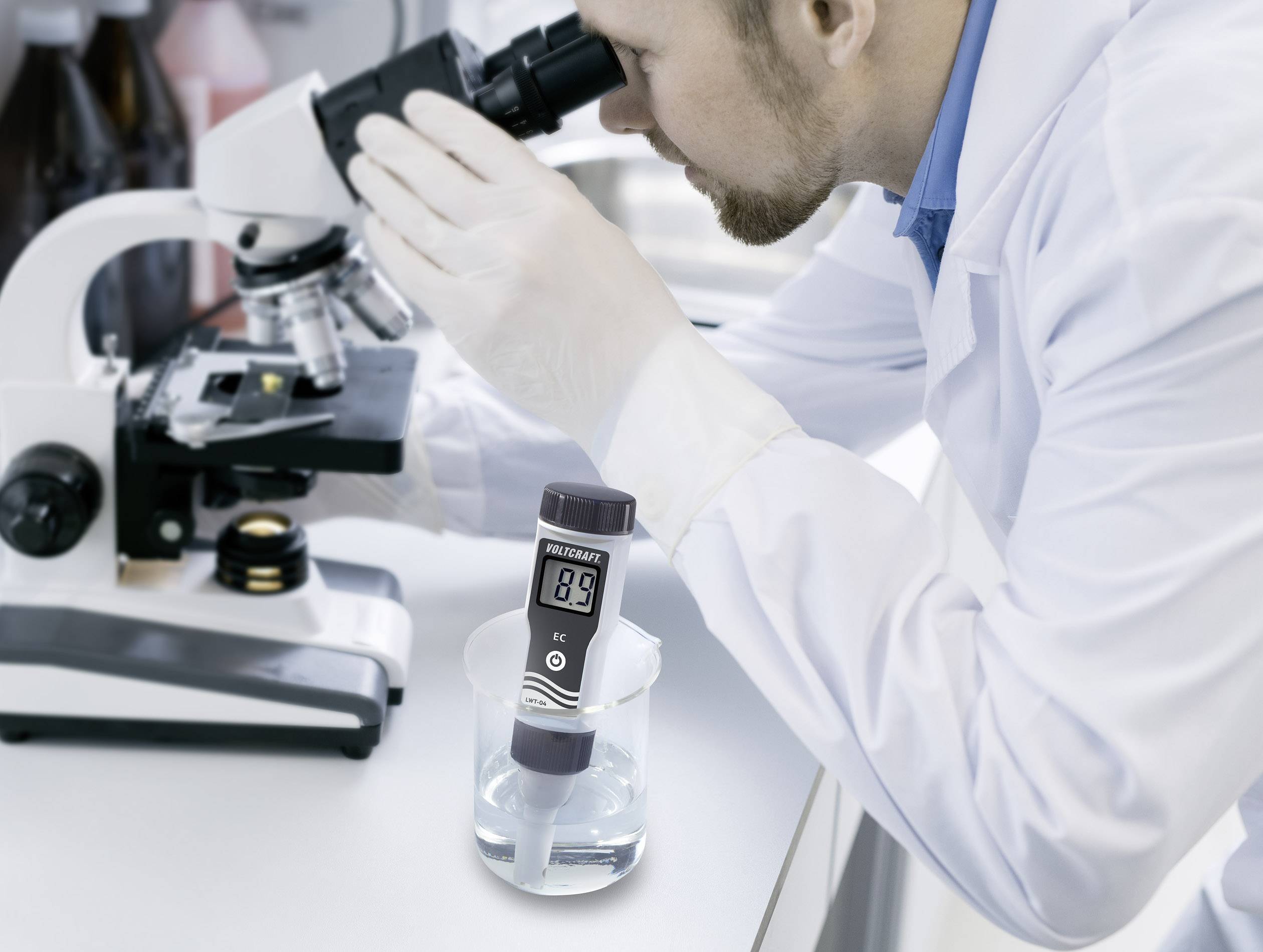 A scientist in a laboratory coat examines samples under a microscope, with a digital measuring device situated beside it in a beaker of liquid.