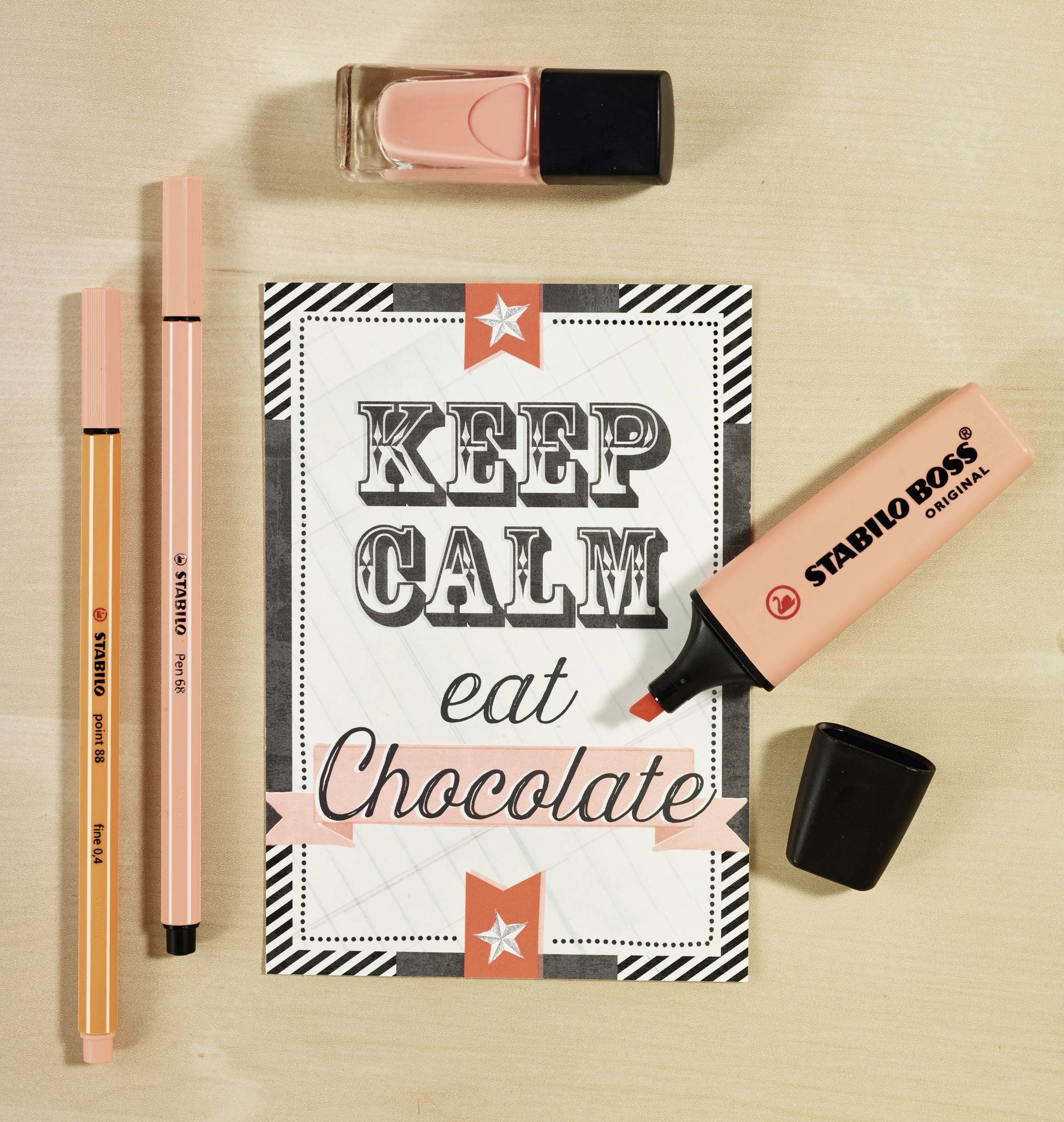 Card with the inscription 'KEEP CALM eat Chocolate' in a decorative design, surrounded by pink pens, markers, and nail polish.