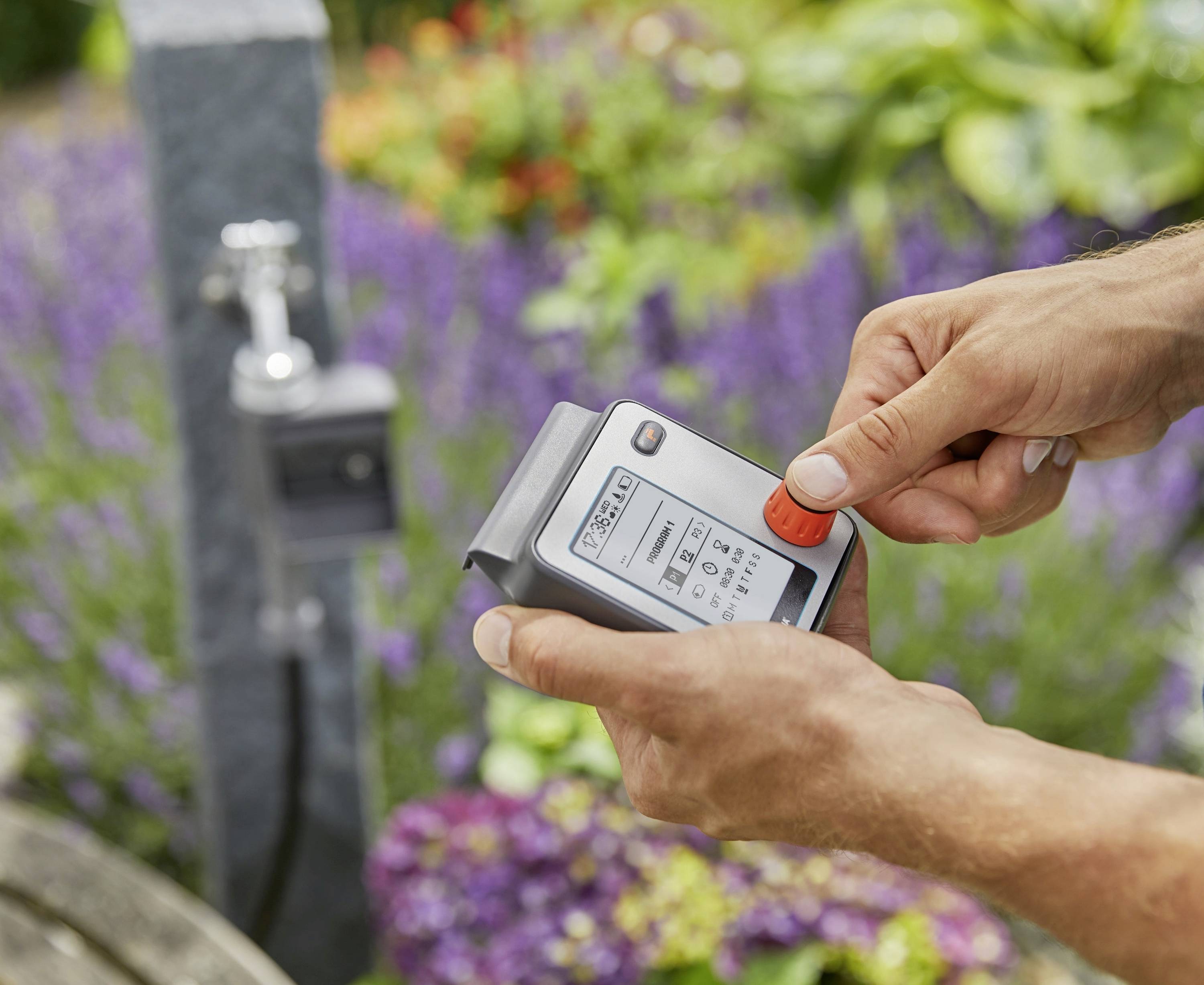 A person is holding a digital irrigation device in front of a garden backdrop with blurred flowers.