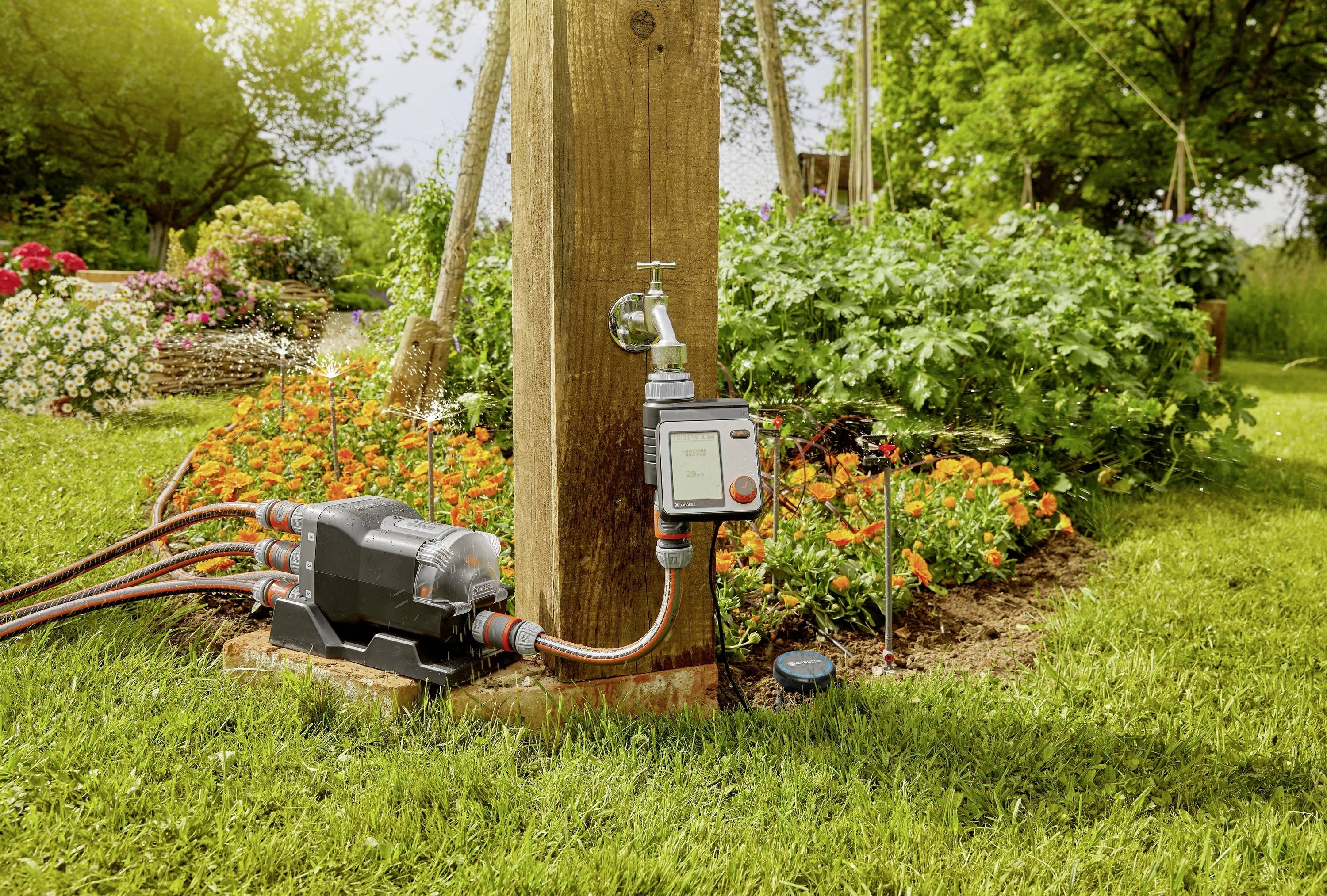 A garden scene with an automatic water pump and timer mounted on a wooden post, surrounded by colourful flower beds.