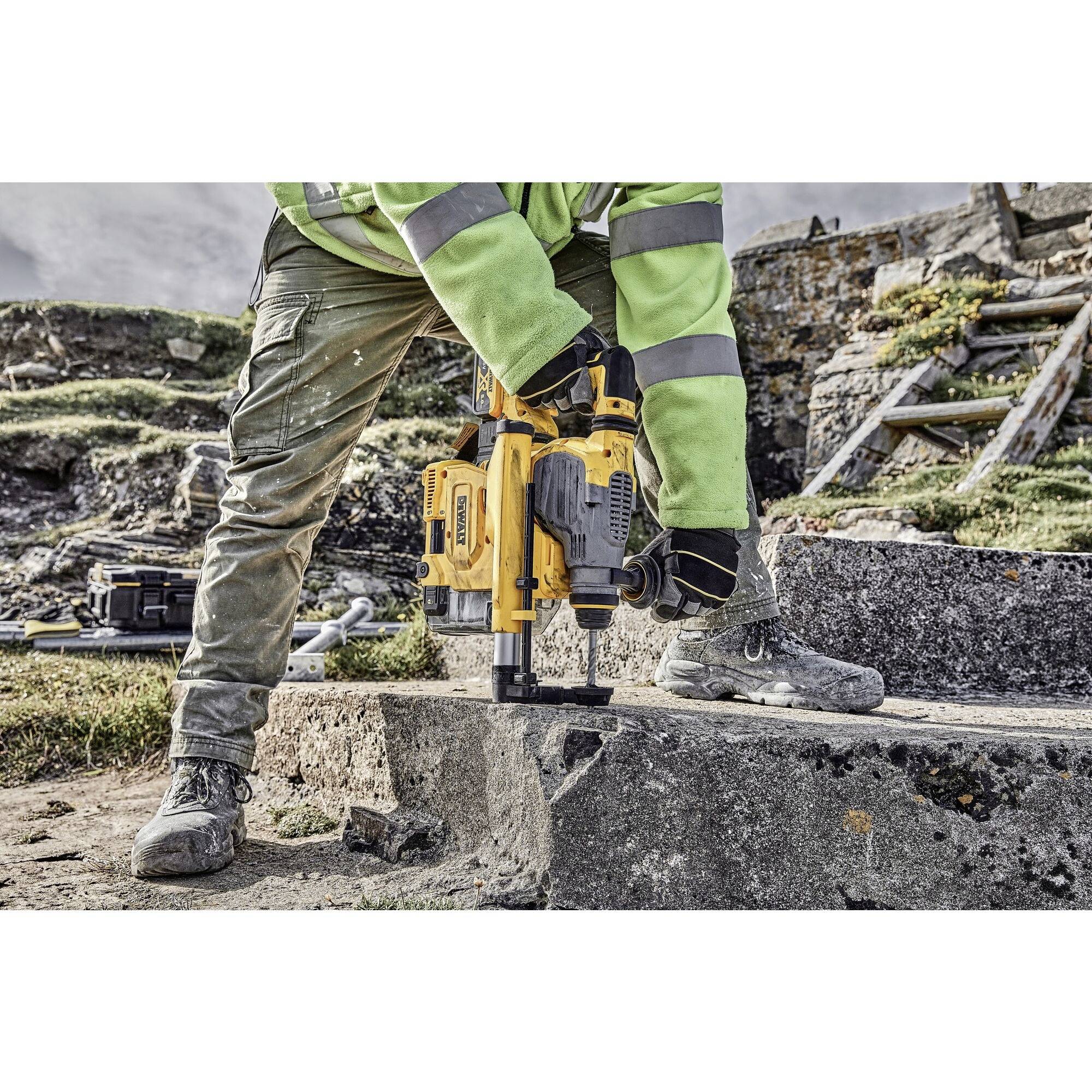 A person in work attire is using a yellow rotary hammer on a stone block in an outdoor construction site environment.