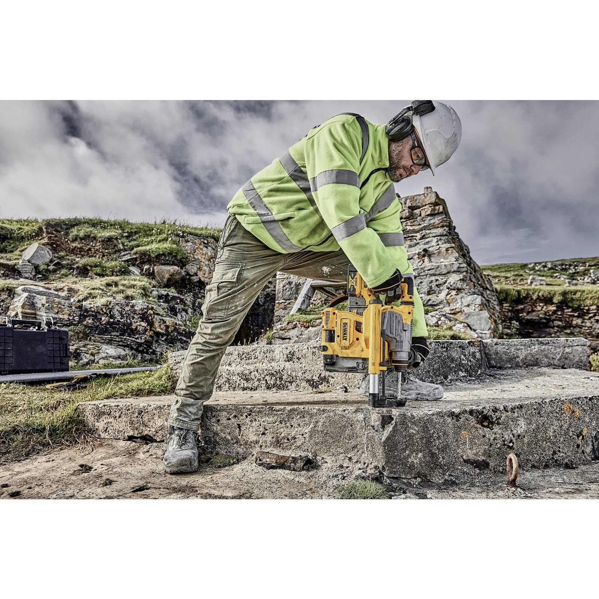 A construction worker in protective clothing is drilling a hole in a concrete block outdoors. Rocks and sky are visible in the background.