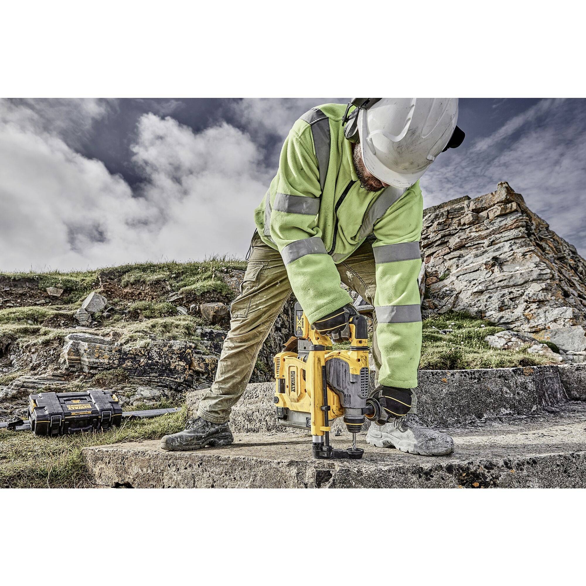 A person in protective clothing is working on a concrete surface with an electric hammer drill in a rocky, cloudy environment.