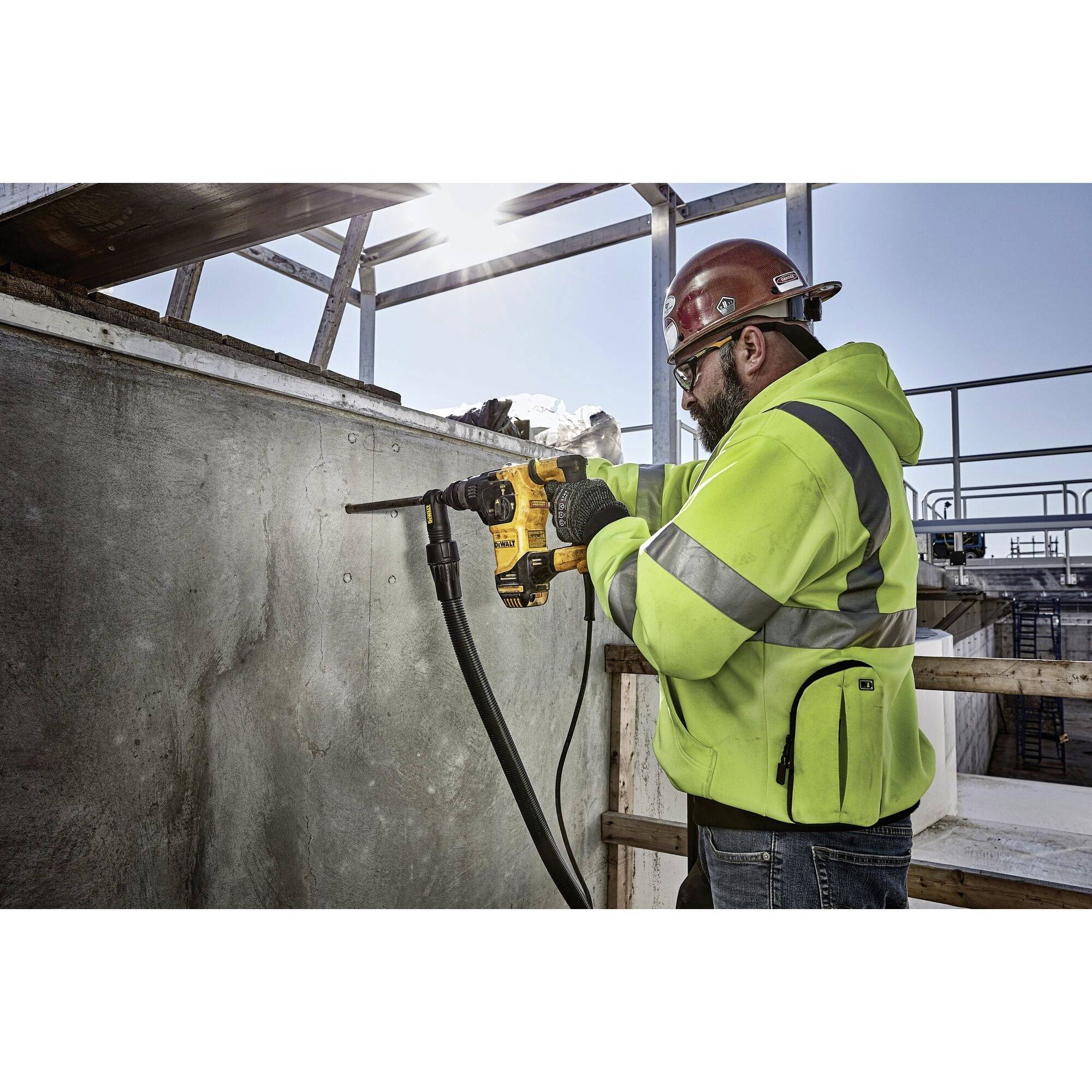 A construction worker in a high-visibility yellow jacket and red hard hat is drilling into a concrete wall on a building site.
