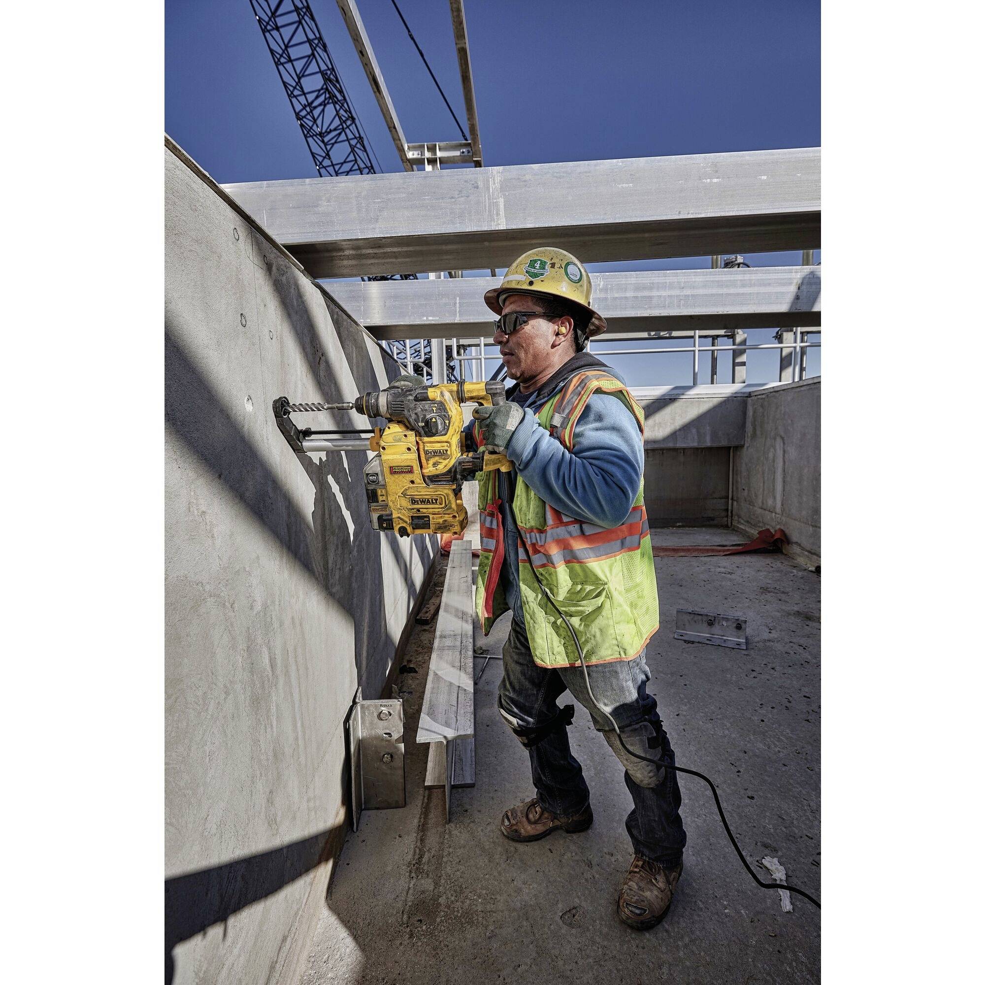 A construction worker wearing a hard hat is using a drill to bore into a concrete wall. A scaffolding is visible in the background.