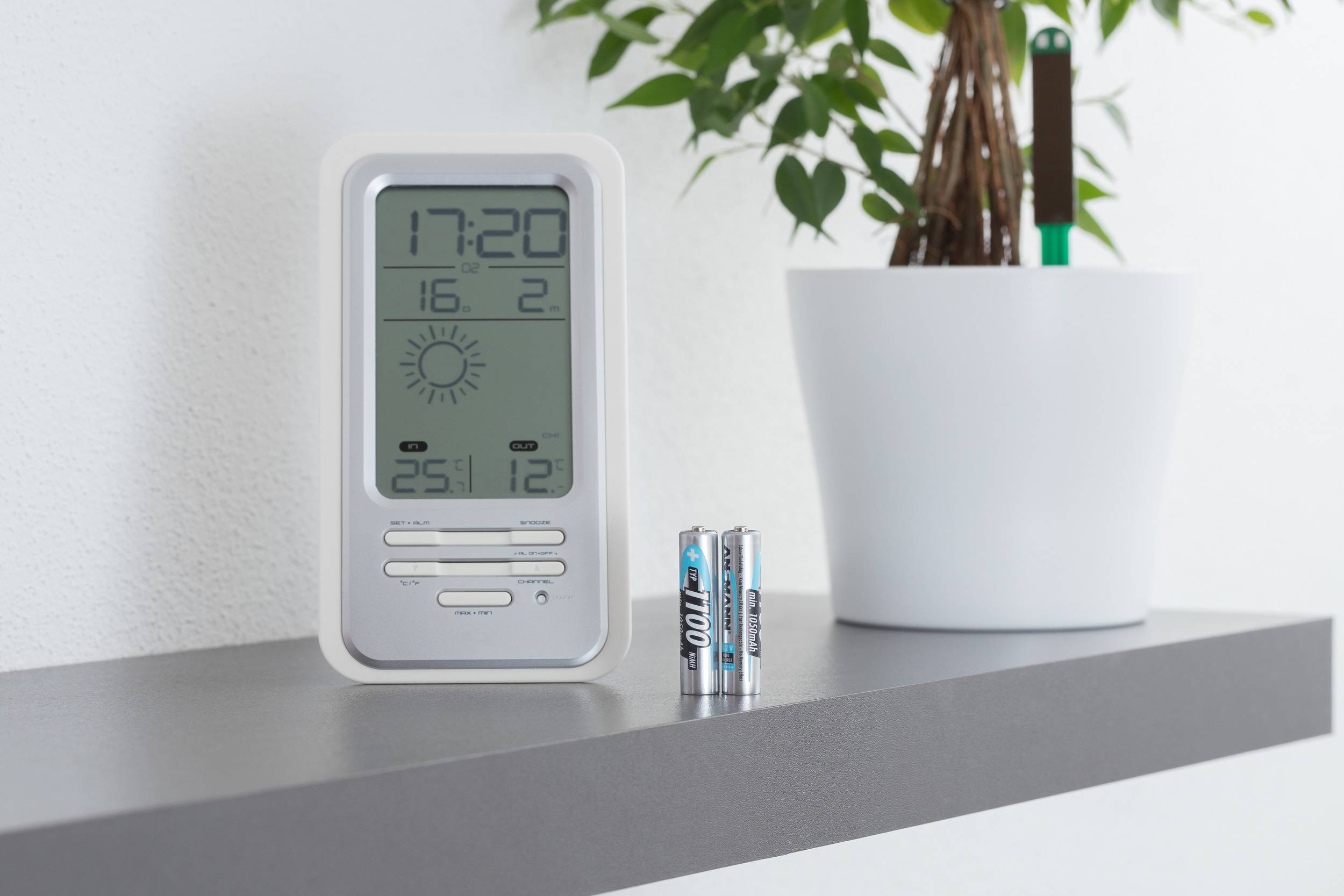 A digital thermometer shows 5:20 pm with temperatures of 25.1°C and 12°C. Two batteries are standing next to it on a shelf, beside a plant.