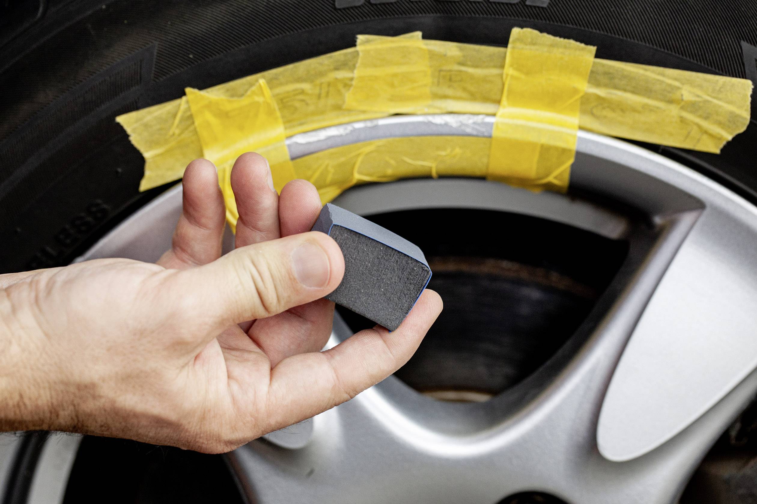 Hand holding sanding block in front of car tyre with yellow-taped rim.
