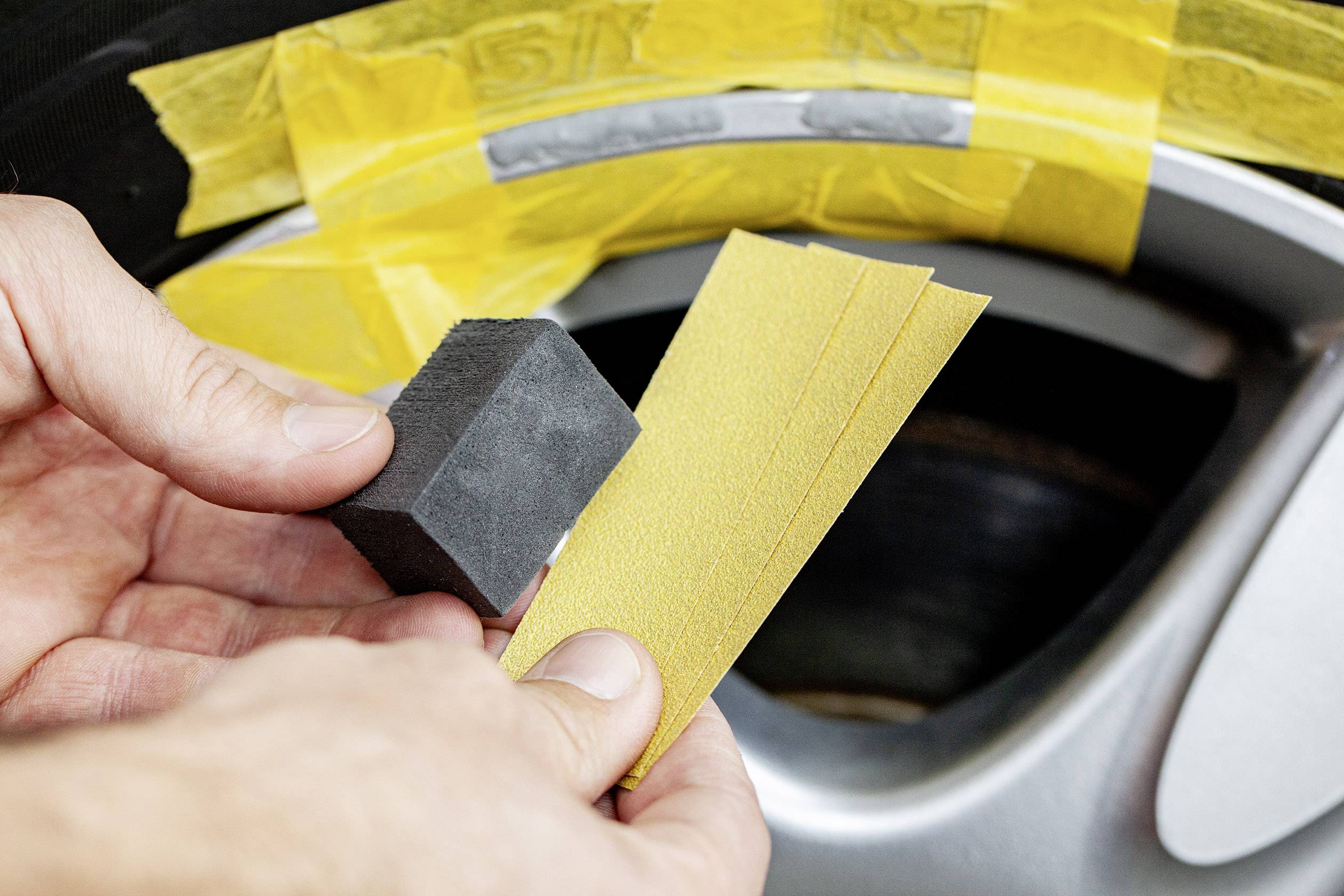 Close-up of hands holding a rubbing tool and sandpaper in front of a car tyre covered with masking tape, ready for processing.