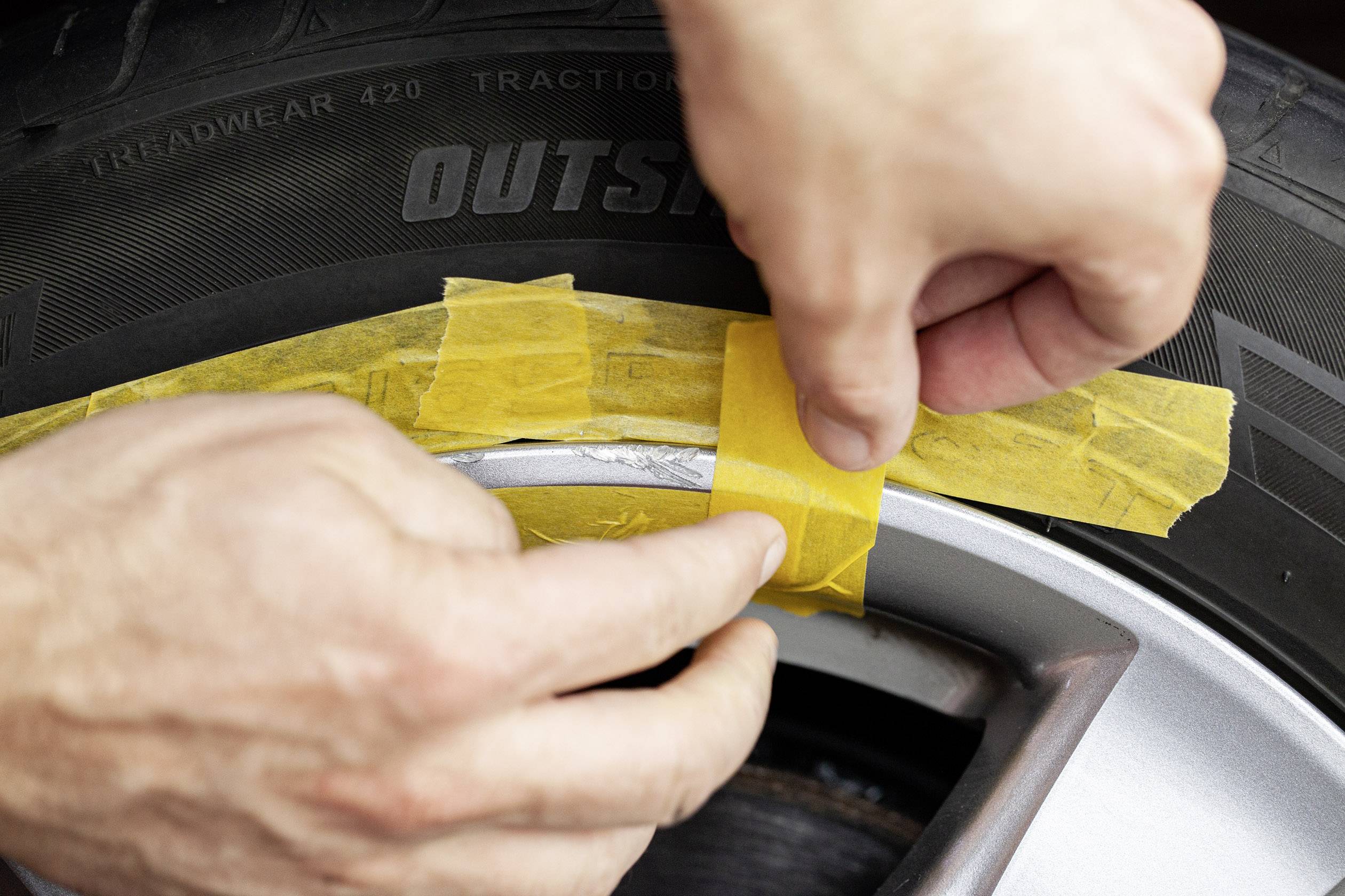 Hands are applying yellow adhesive tape to the rim of a car tyre. The tape protects the rim from damage when working on the tyre.