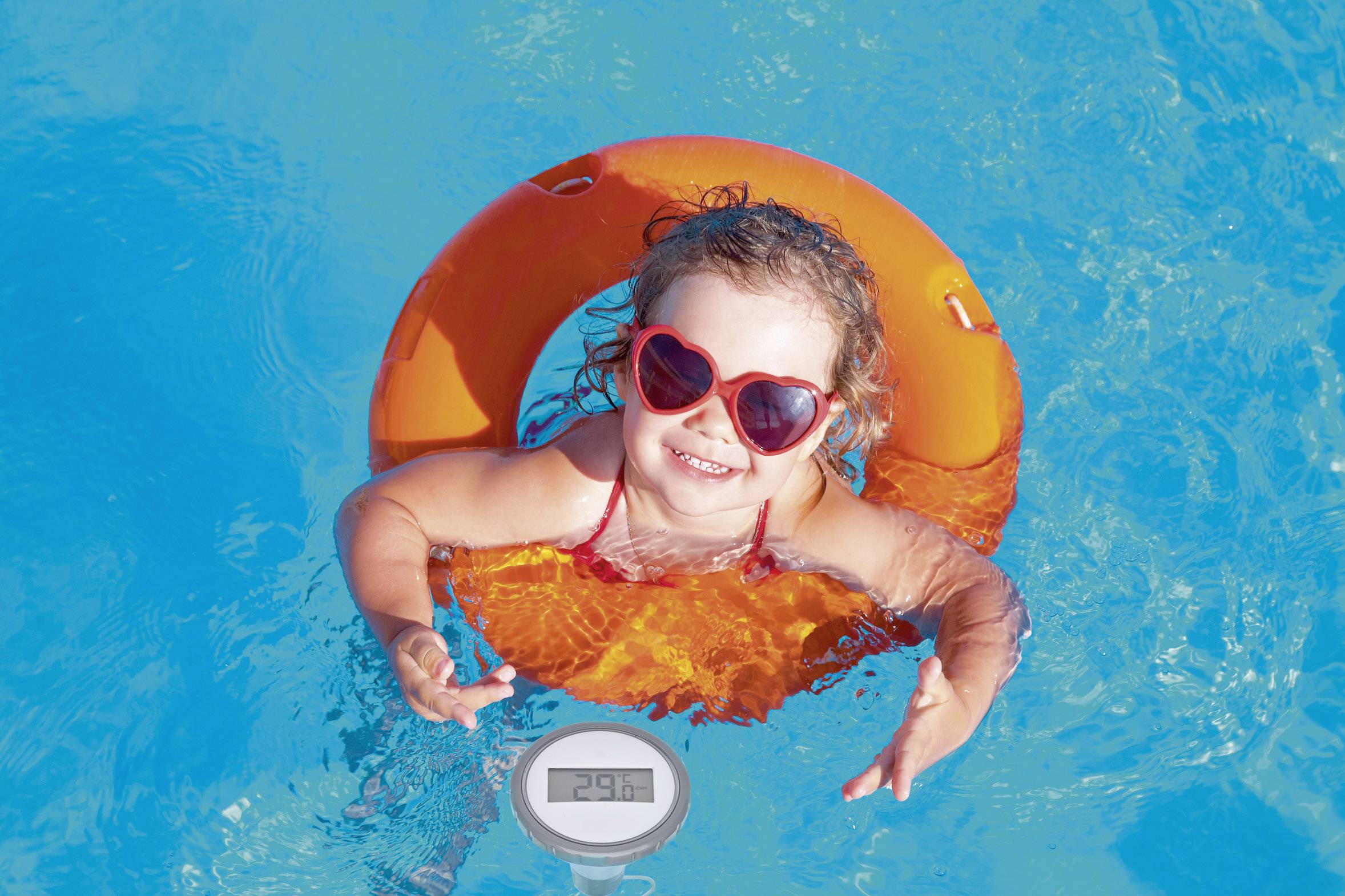 A smiling little girl with heart-shaped sunglasses swims in the blue pool on an orange rubber ring.