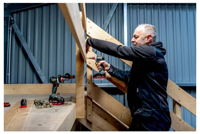 Man using a cordless drill to fasten wooden beams on an indoor staircase construction project.