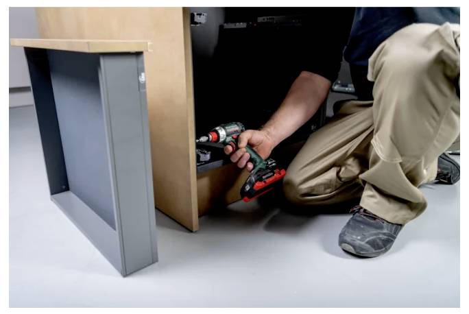 Person kneels under a cabinet, using a cordless drill to install or repair furniture.