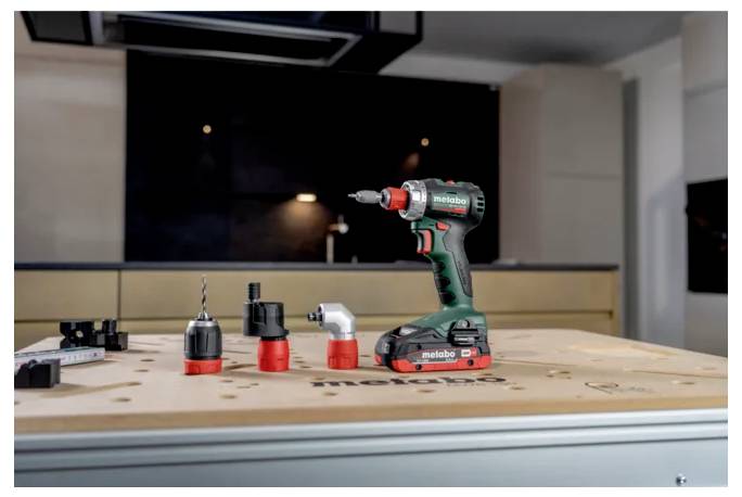 Cordless Metabo drill standing on a workbench with three drill attachments in a workshop kitchen setting.