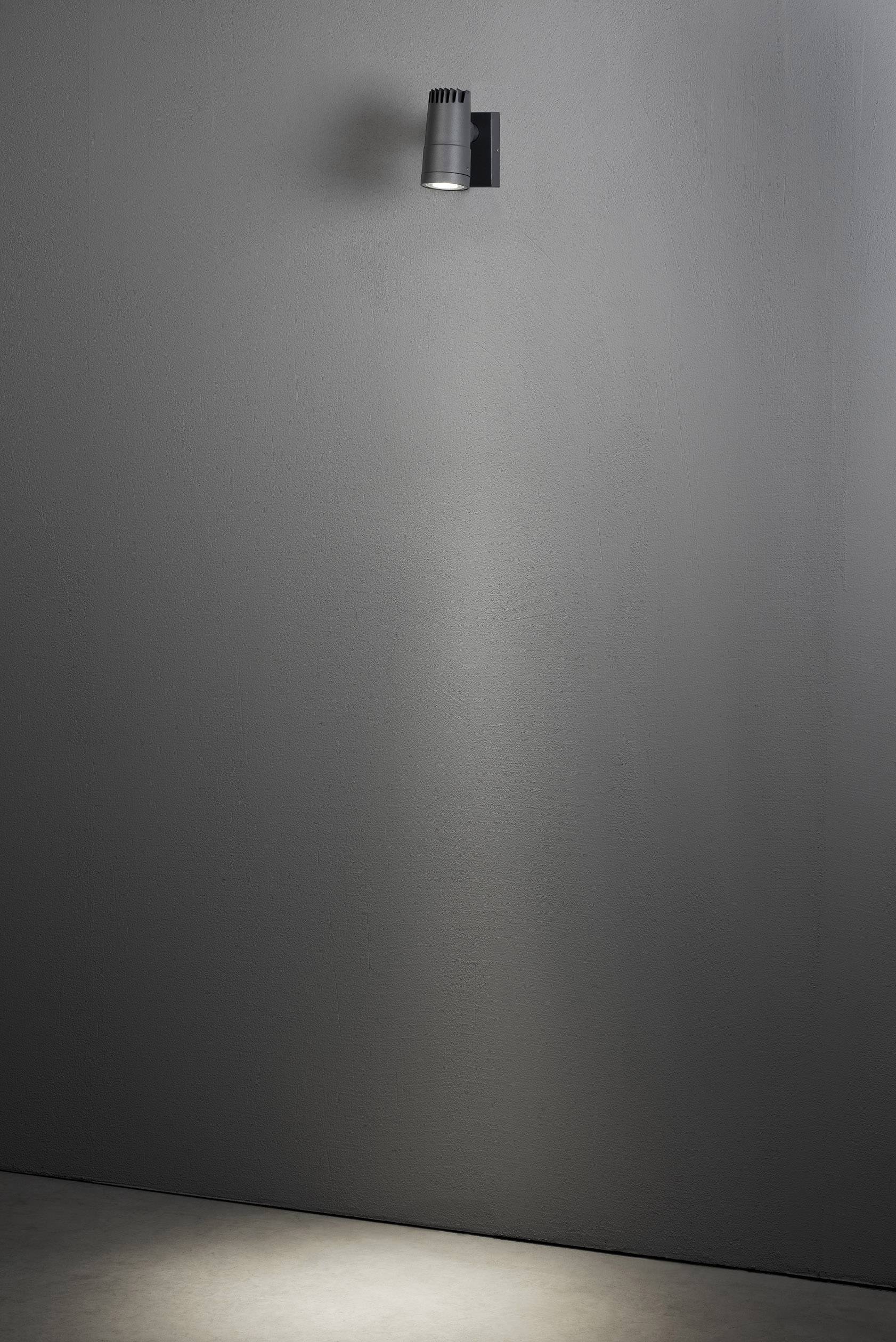 A single wall light illuminates an area on an otherwise bare grey wall, creating a bright cone of light.