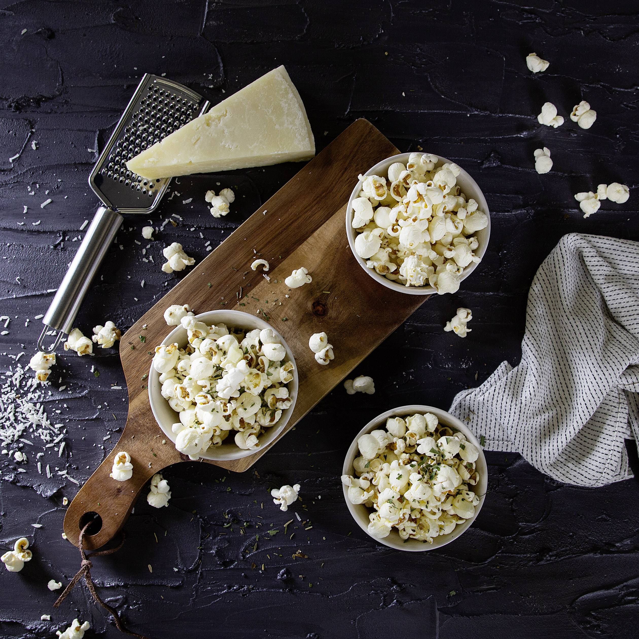 Three bowls of popcorn on a wooden chopping board, surrounded by grated cheese, a cheese grater, and a tea towel.