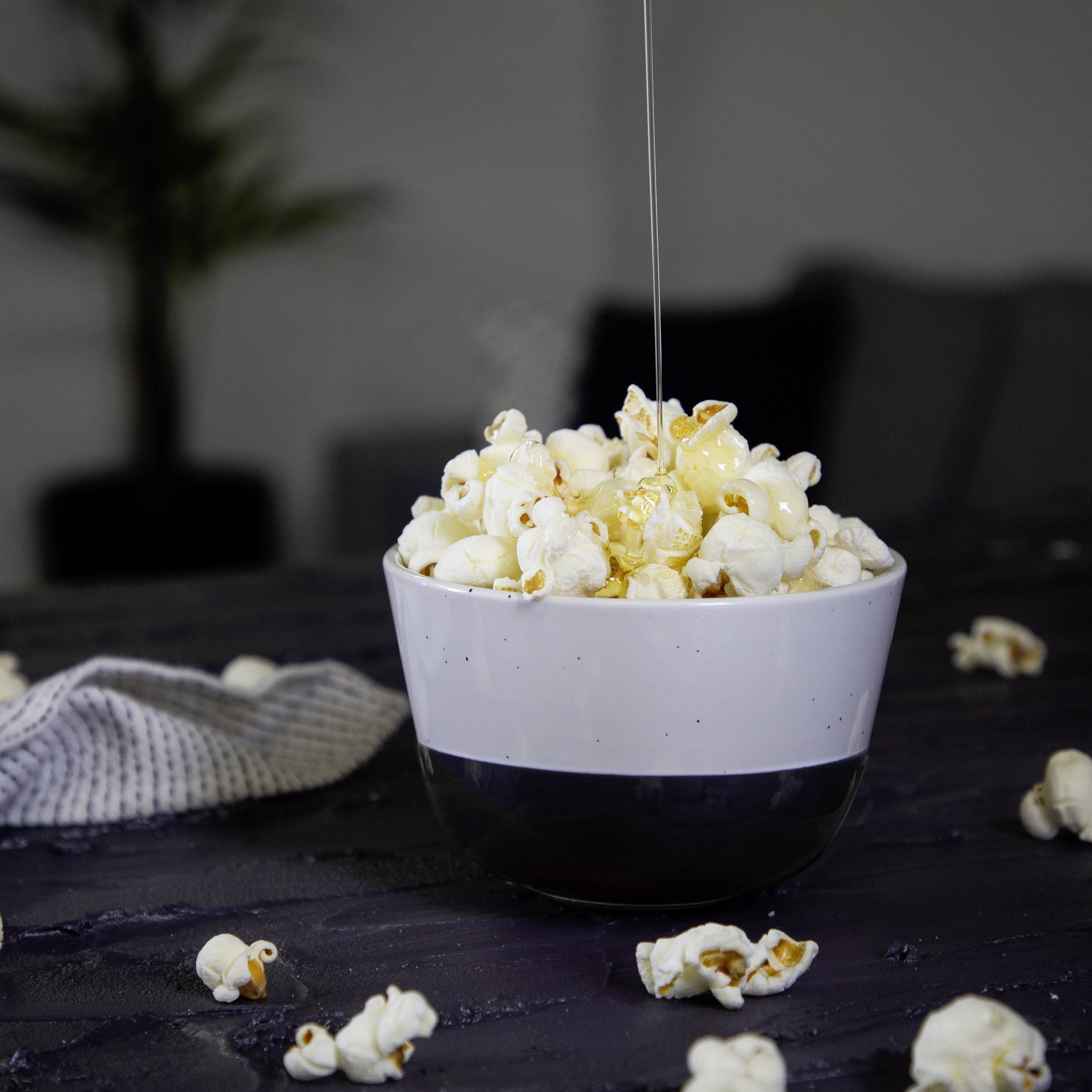 A bowl of popcorn on a table, with honey being poured over it, against a blurred background.