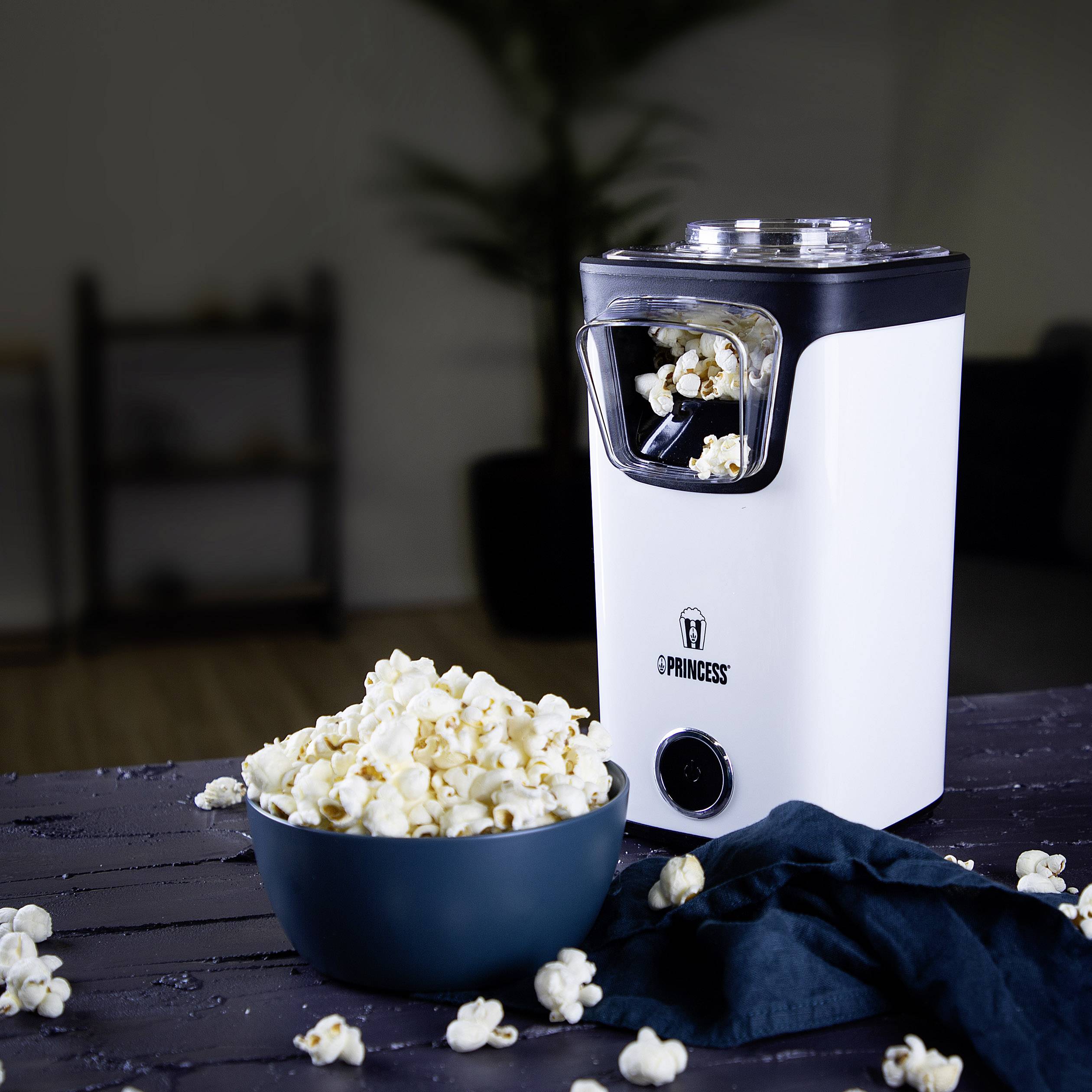 A white popcorn machine with a bowl full of popcorn beside it. Blurred shelves and plants are visible in the background.