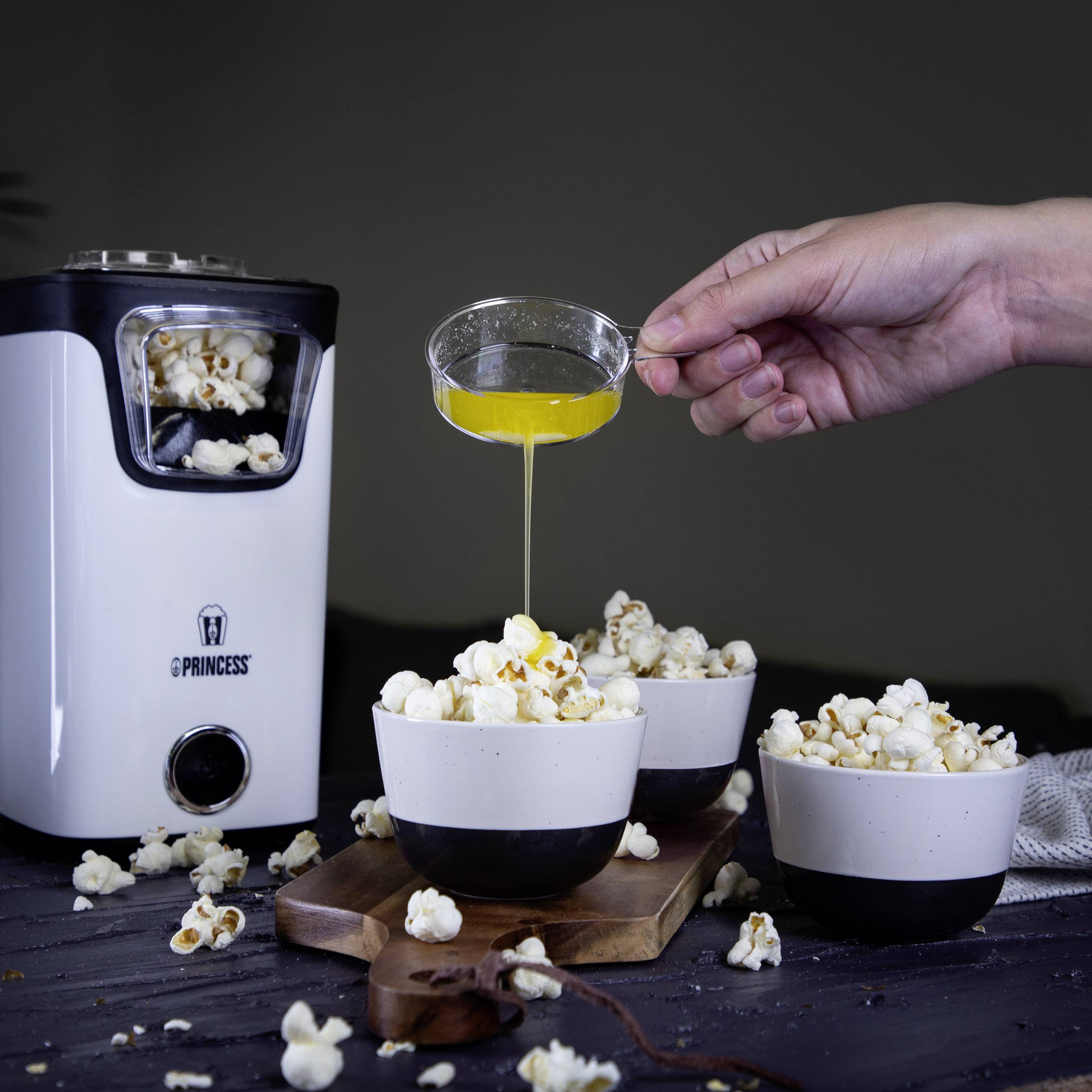 Popcorn machine fills bowls with popcorn. A hand pours melted butter over the popcorn. Dark background.