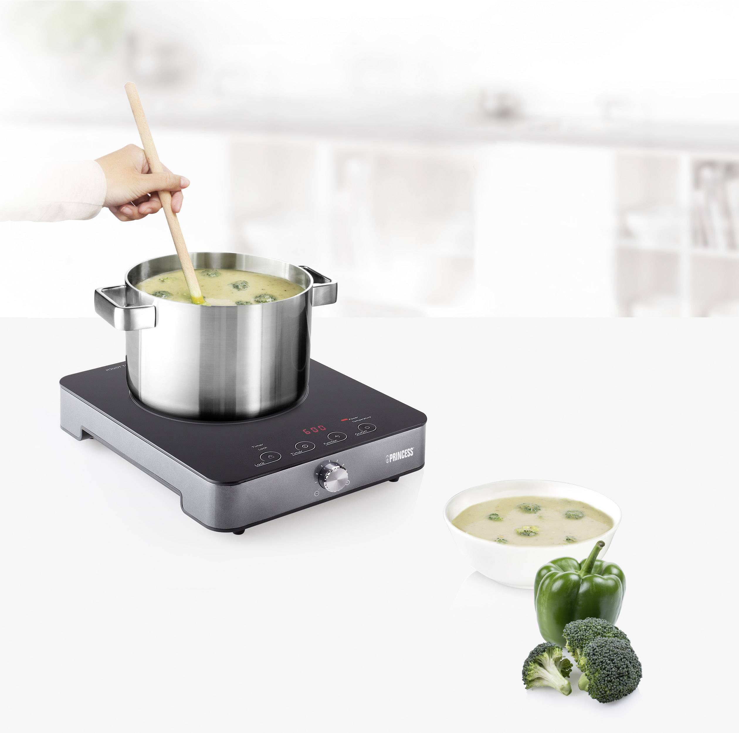 A hand stirs a pot of soup on a hob. Beside it is a bowl of soup with broccoli and a green pepper.