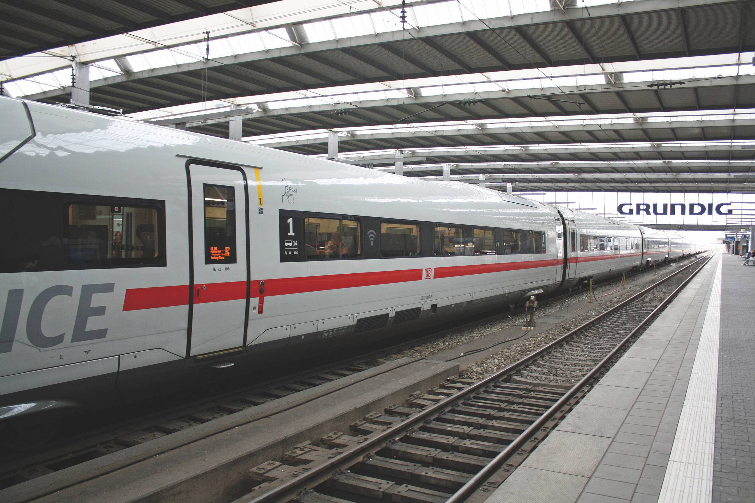 A modern ICE train stands on a platform in a covered railway station. The word 'ICE' is written on the train.