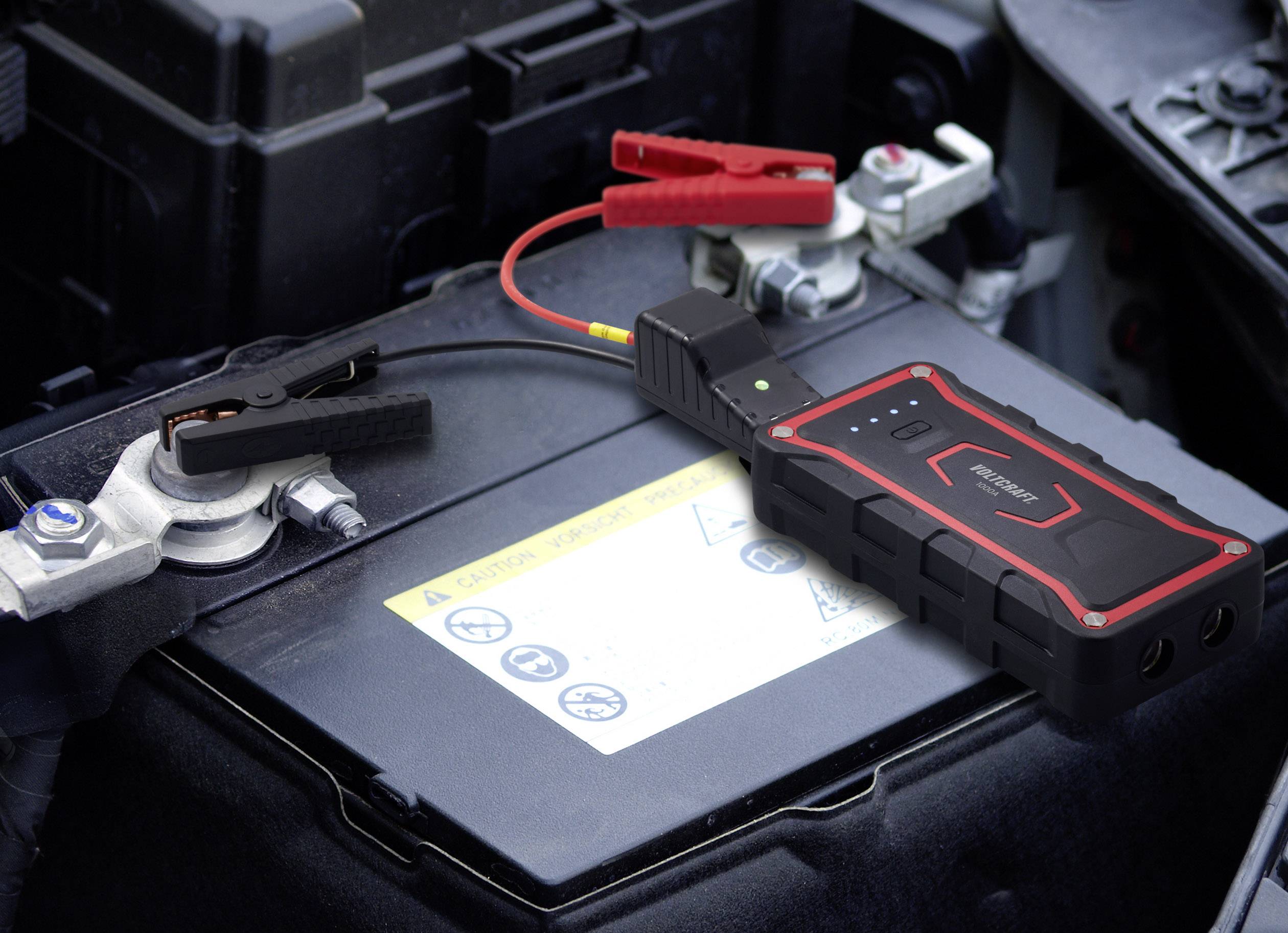 A car battery is being charged by a mobile jump starter. The clamps are correctly attached to the battery terminals.