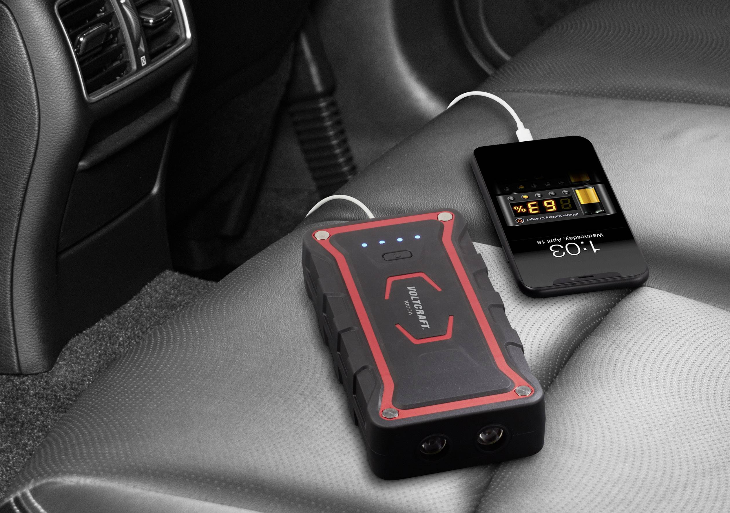 A portable battery pack is resting on a car seat, charging a smartphone via a USB cable. The phone's display shows charging information.