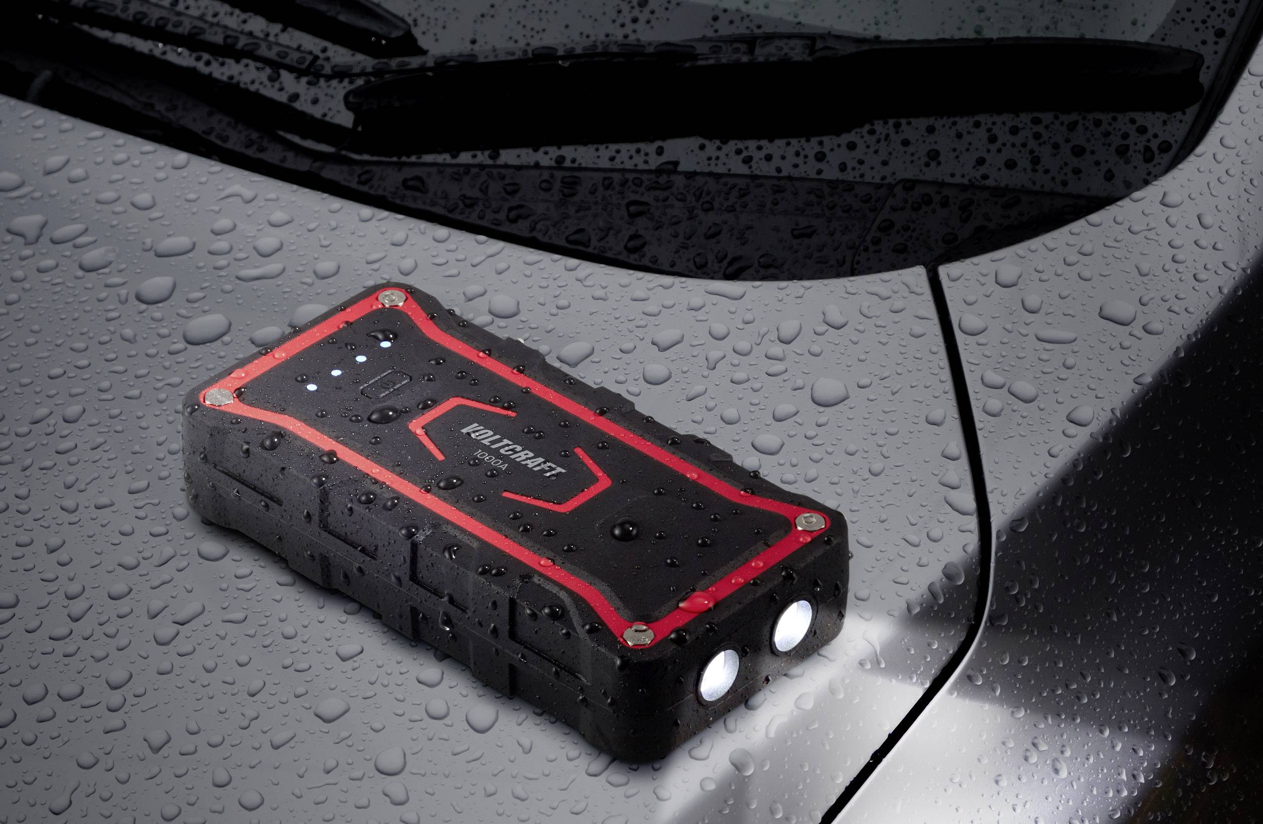 A black, portable car battery jump starter with red accents and LED lights is lying on the wet bonnet of a car.