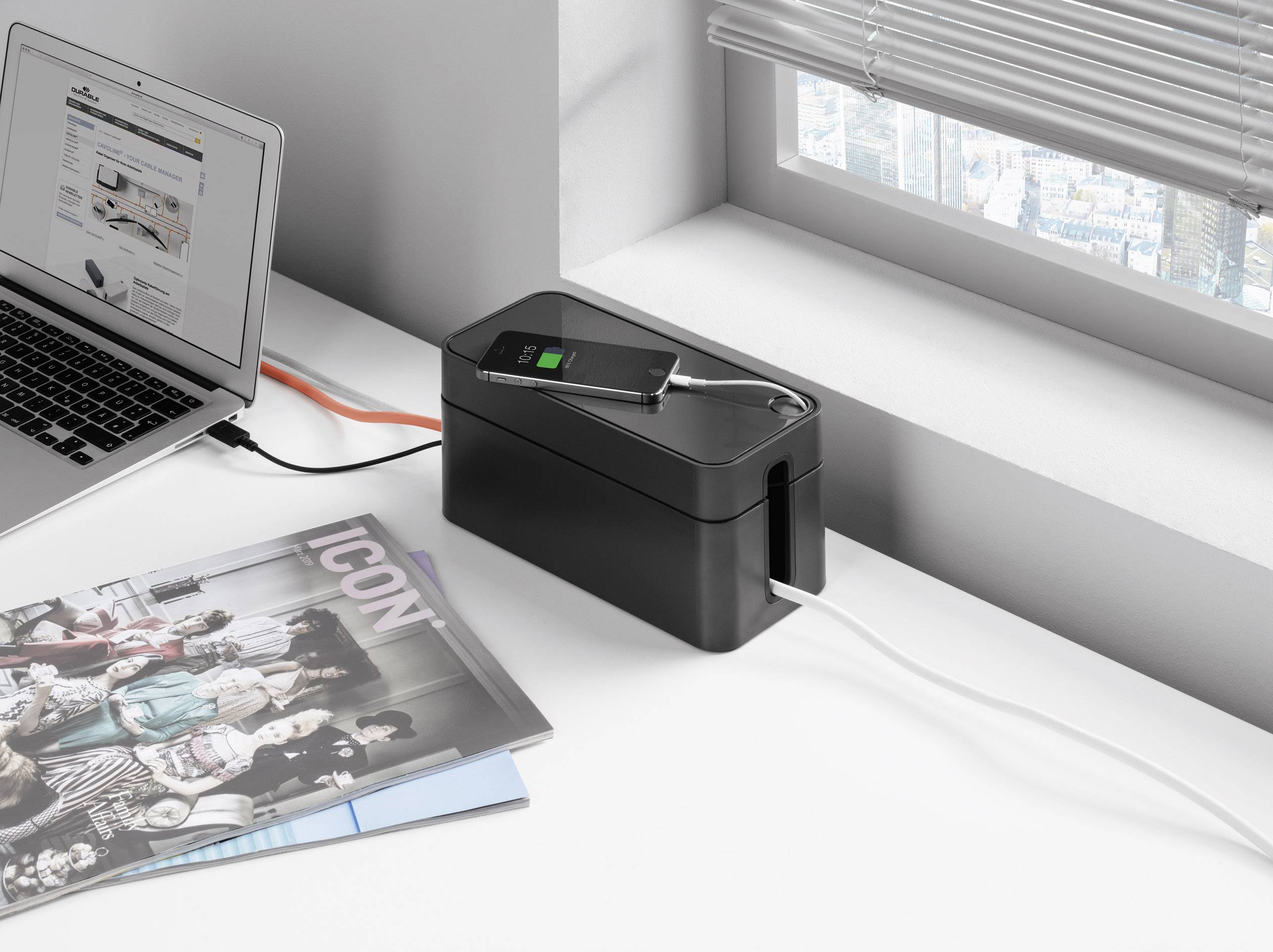 A laptop is connected to a cable management box. A smartphone is charging on the box. A magazine is lying next to the box.