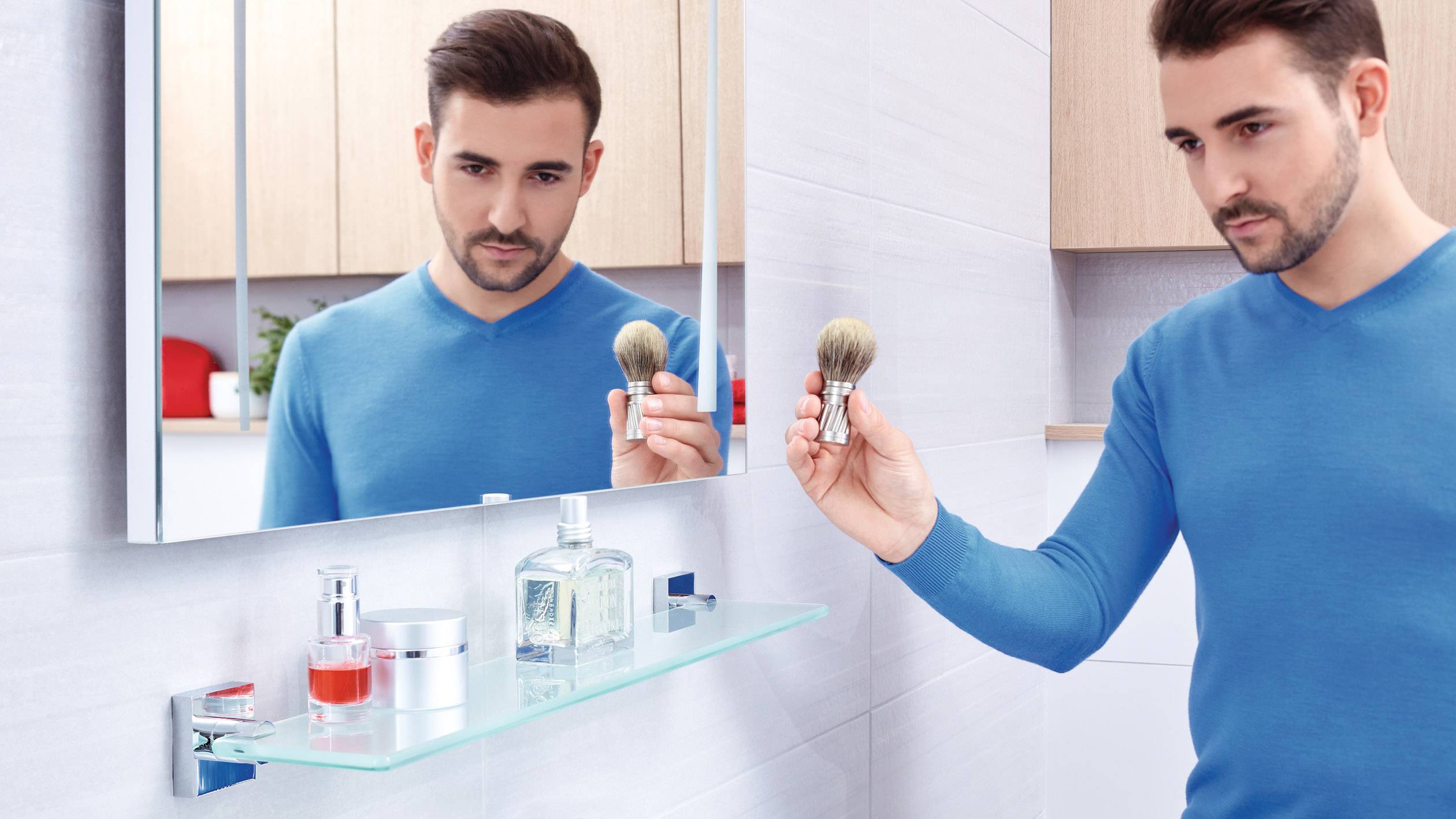 A man in a blue jumper looks at himself in the mirror while holding a shaving brush. Cosmetic items are placed on a glass shelf in the foreground.