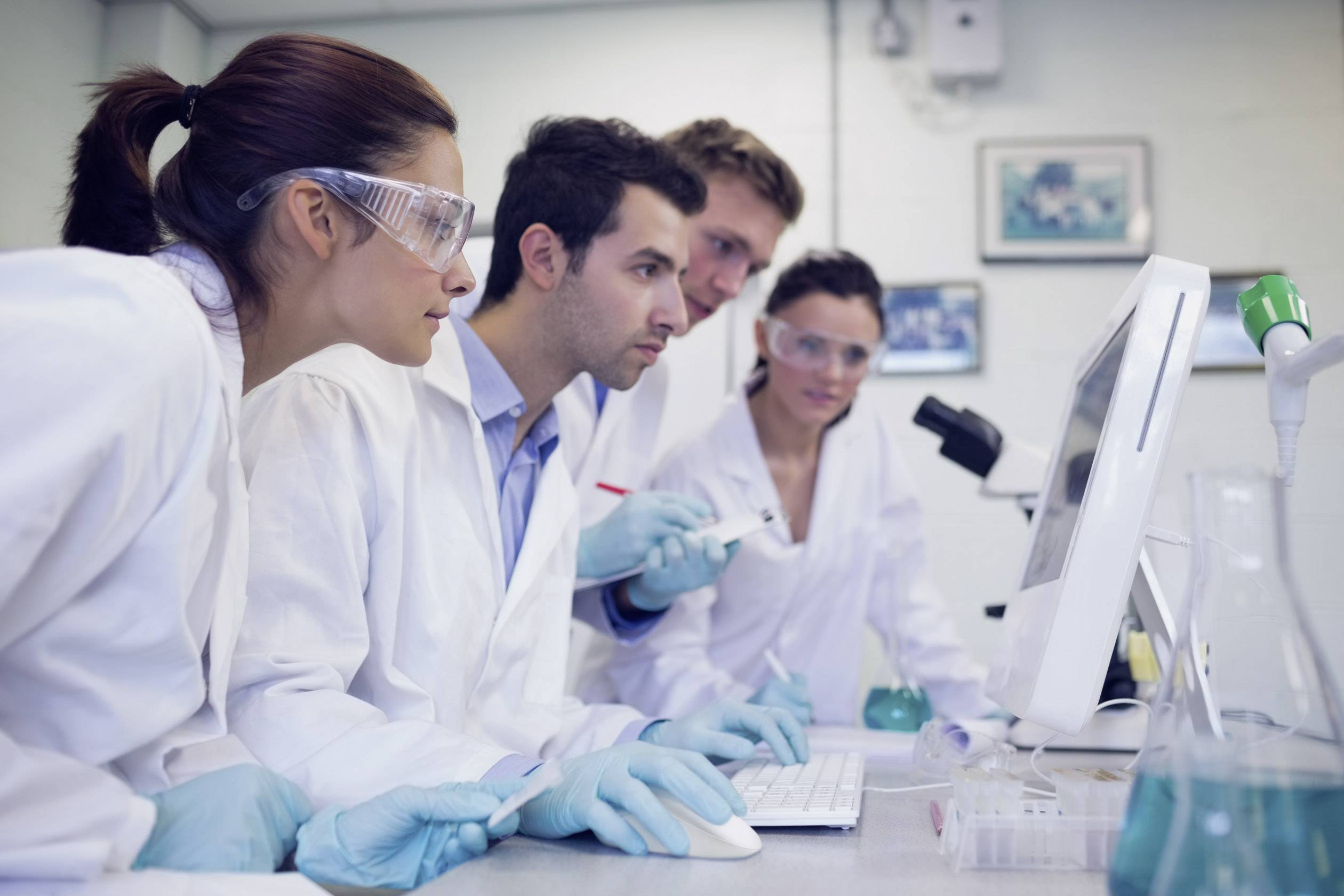 Four individuals in lab coats and safety glasses are working intently at a computer in a laboratory, surrounded by scientific equipment.