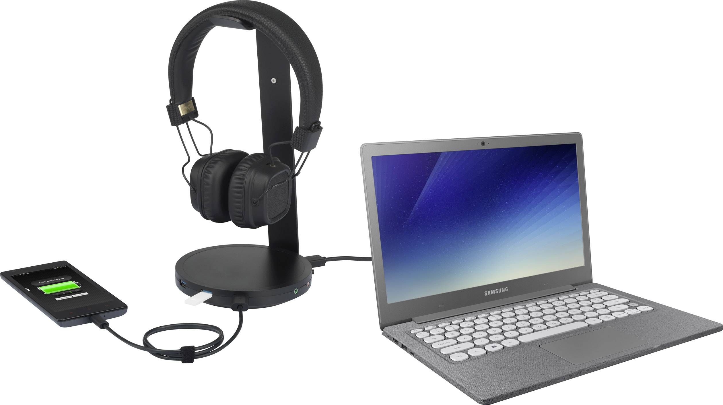 A laptop, a smartphone with a charging indicator, and headphones on a stand are arranged side by side. The laptop is switched on.