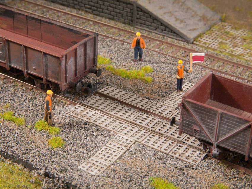 Workers in orange hi-vis vests stand on a model railway site next to two freight wagons on the tracks. One is holding a flag.
