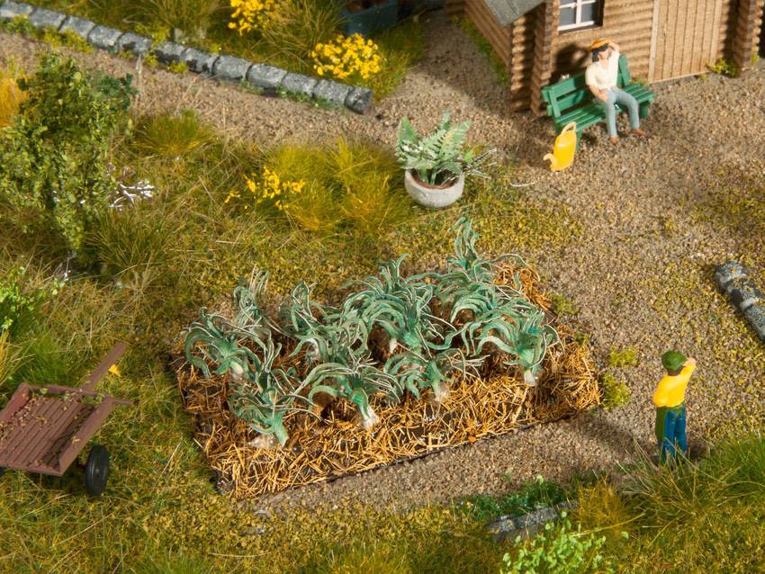 A miniature scenario depicts a vegetable garden with plants on a straw bed. Two figures can be seen in a relaxed setting.