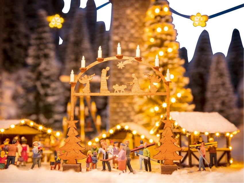 A illuminated wooden Christmas arch with figurines, decorated huts and trees in the background, creates a festive atmosphere.