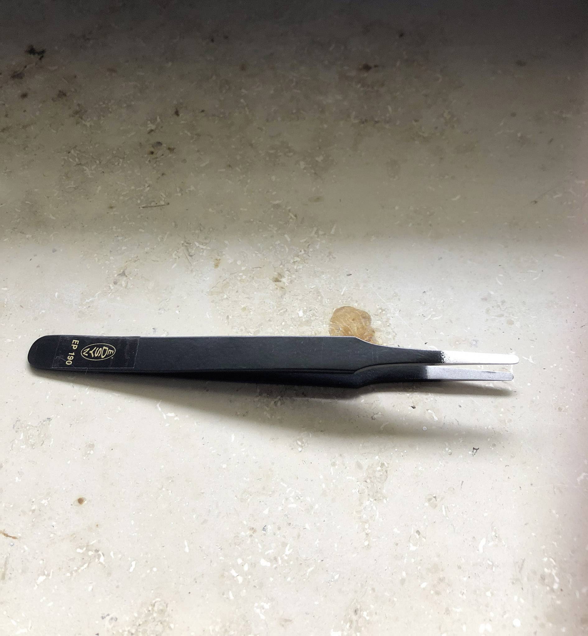A black pair of tweezers with a white insert is resting on a light, textured surface.