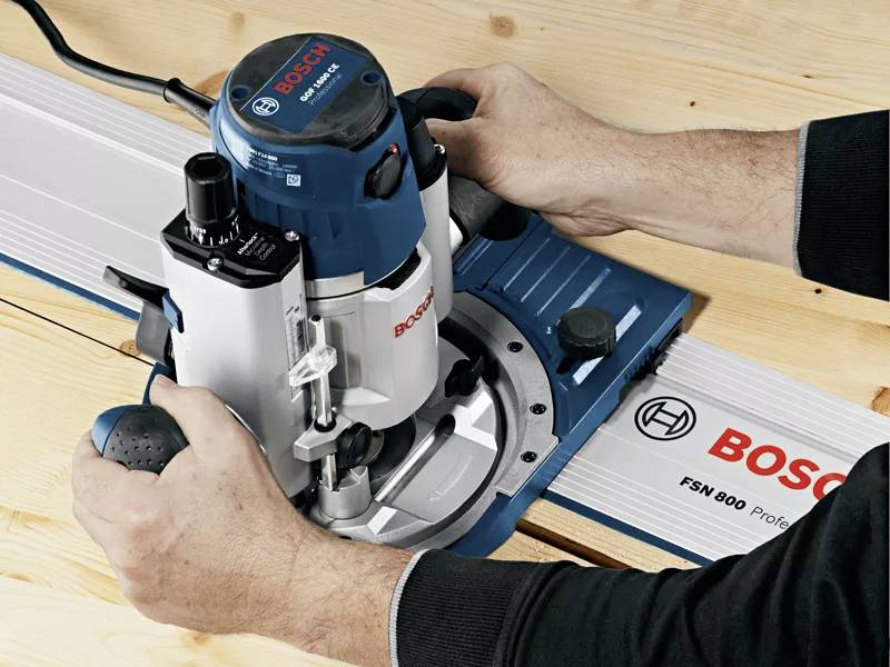 Two hands guide a blue Bosch router along a guide rail on a wooden table.