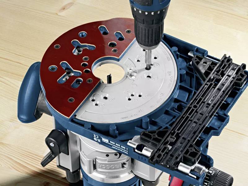 An electric milling cutter drills into a circular metal plate. The milling machine is positioned on a light-coloured wooden table.