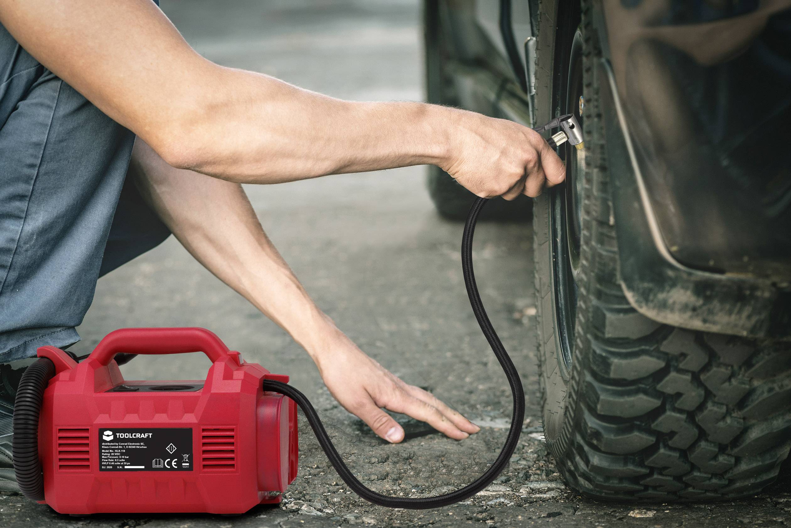 A person is inflating a car tyre with a red air compressor. The compressor is standing next to the vehicle on the ground.