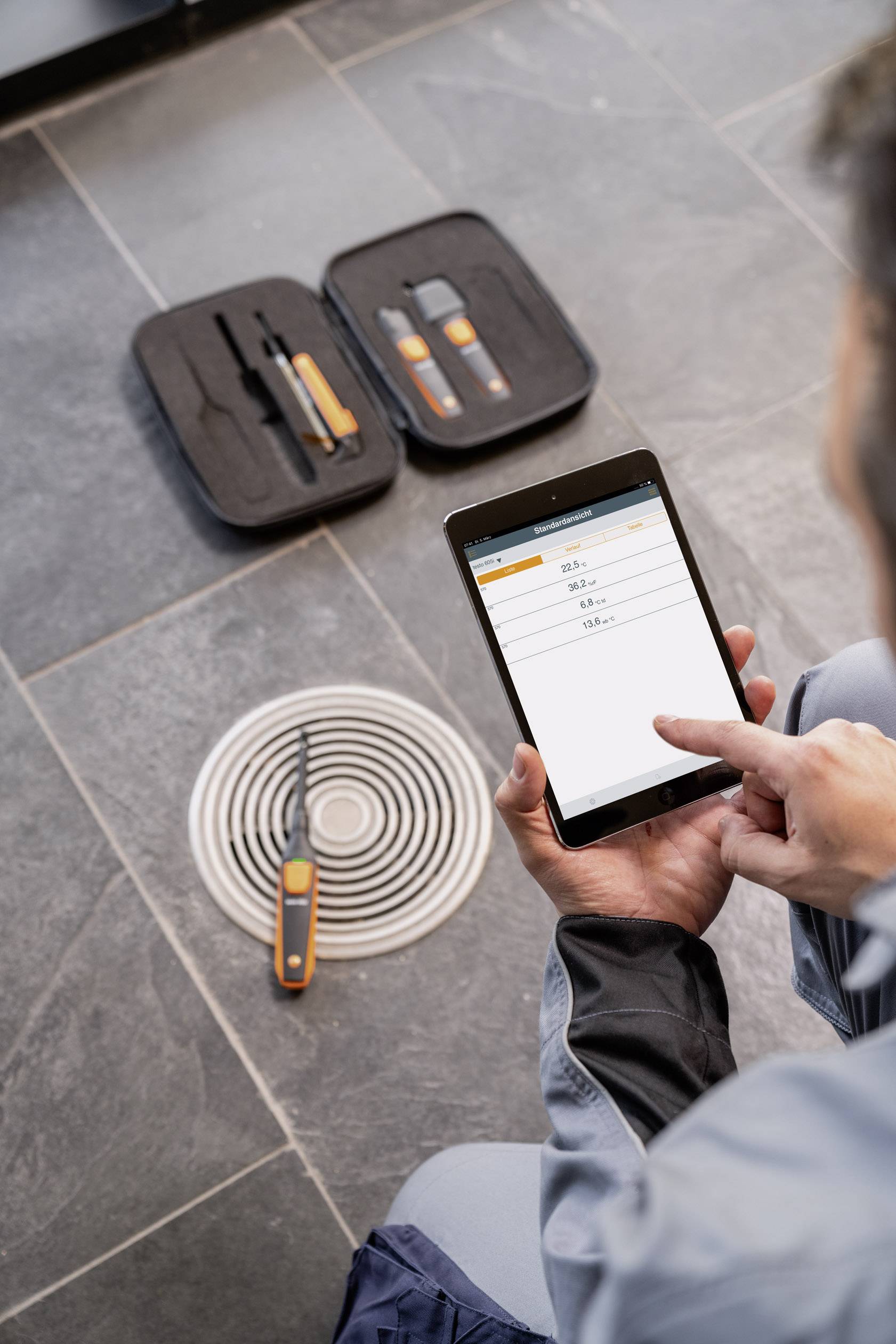 A person is operating a tablet to measure data, with tools lying beside them on a floor drain.