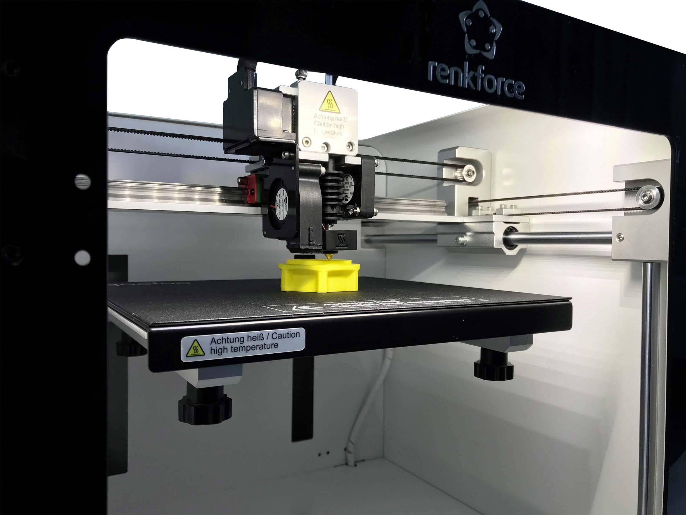 A 3D printer is printing a yellow object. Warning sign on the platform: 'Caution - Hot Surface / Caution High Temperature'.