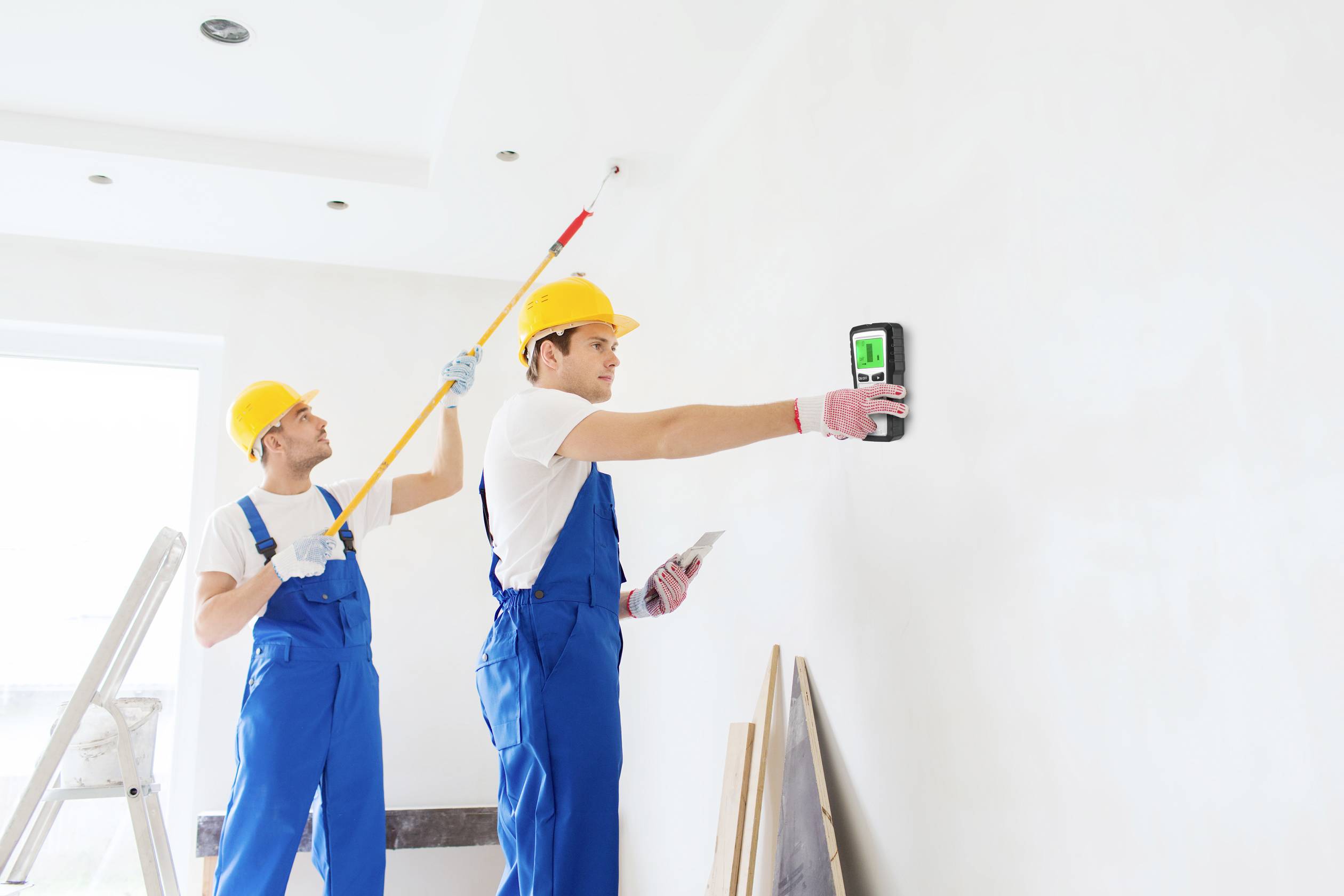 Two construction workers in blue overalls and yellow hard hats are painting a white wall. One is measuring the wall while the other is painting.