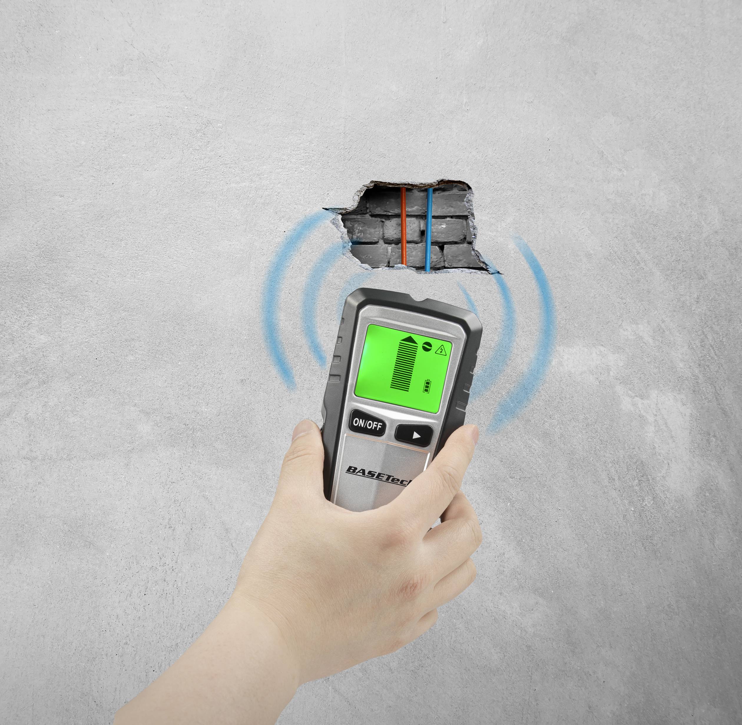 A hand holds a digital locator device against a wall with a hole, behind which are red and blue pipes. The device is showing location waves.